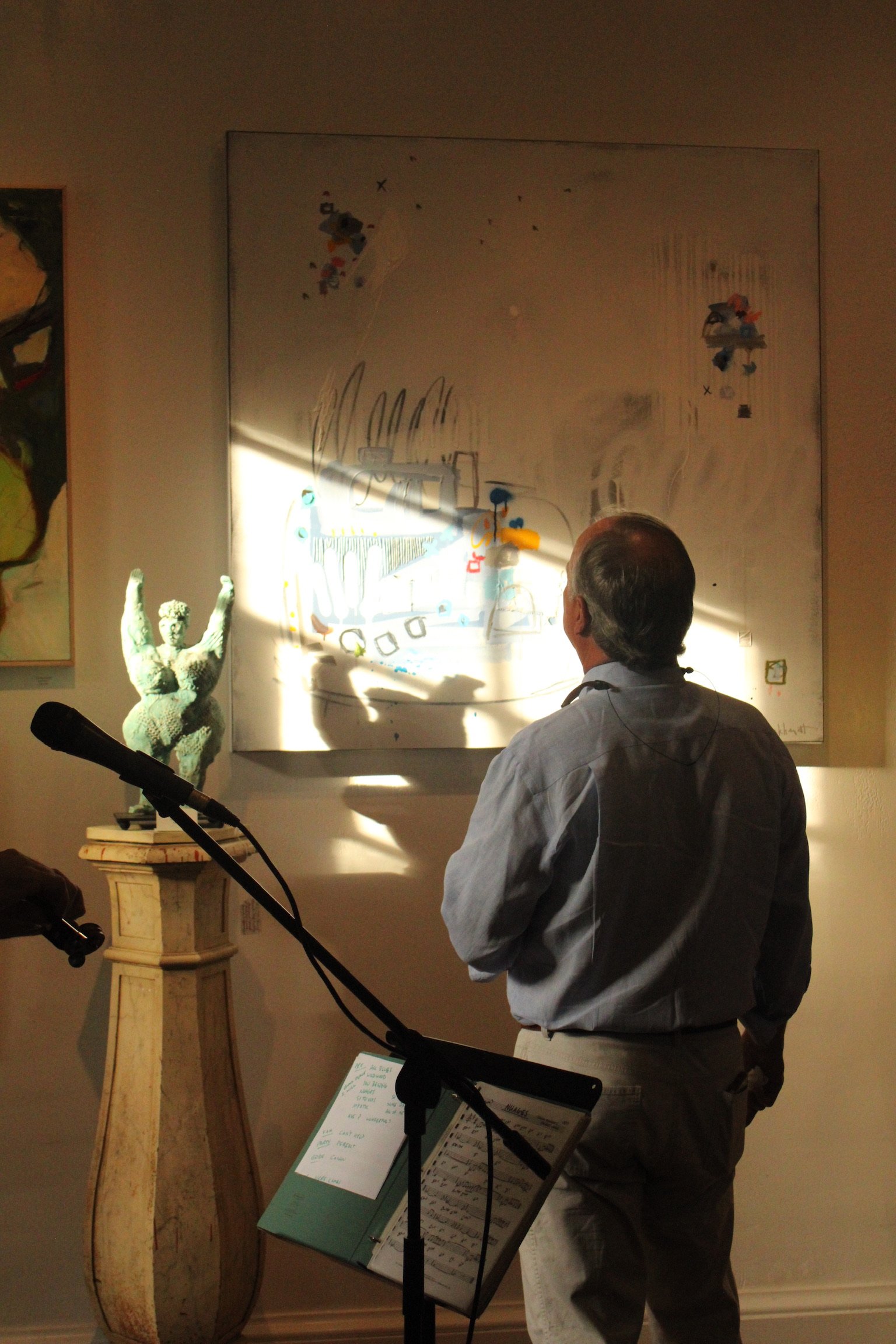 A man with gray hair, wearing a light blue shirt and glasses, stands in front of an art exhibit in a gallery. He is looking at a large white canvas with abstract drawings. In front of him is a music stand with sheet music, and there is a statue of a 