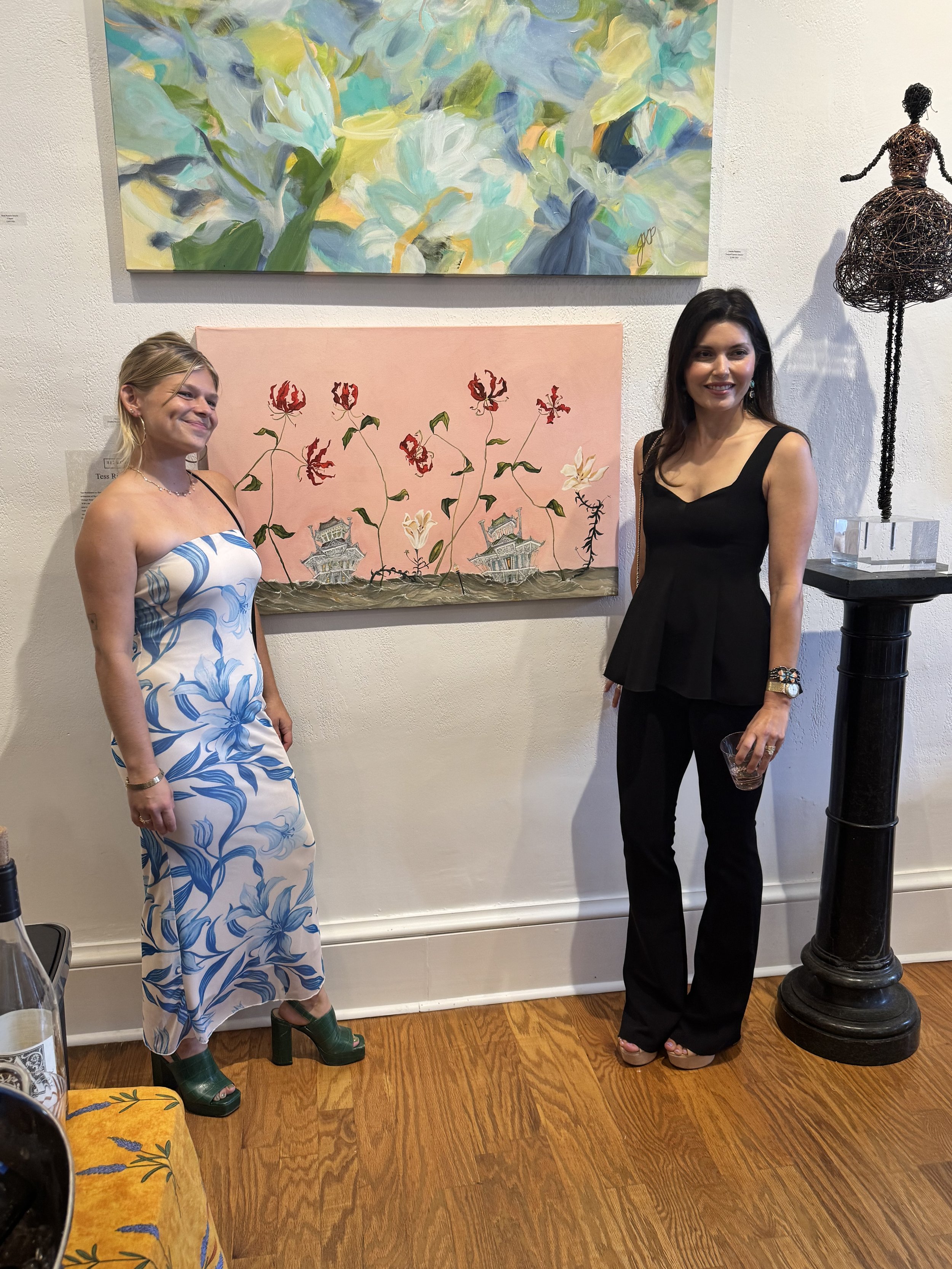 Two women standing in an art gallery, smiling. Behind them are two paintings on the wall: the top one is an abstract painting with blues, greens, and yellows, and the bottom one features red and white flowers with small houses. To the right is a wire