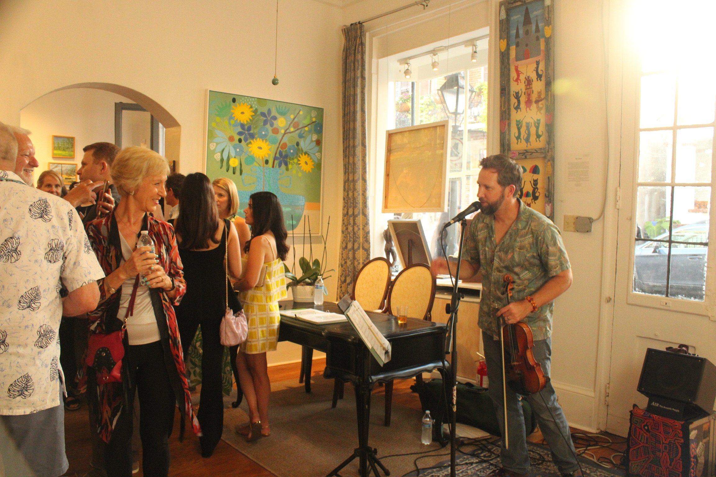 A man playing guitar and singing into a microphone at a gathering or event, surrounded by several people in a room with colorful artwork and large windows.