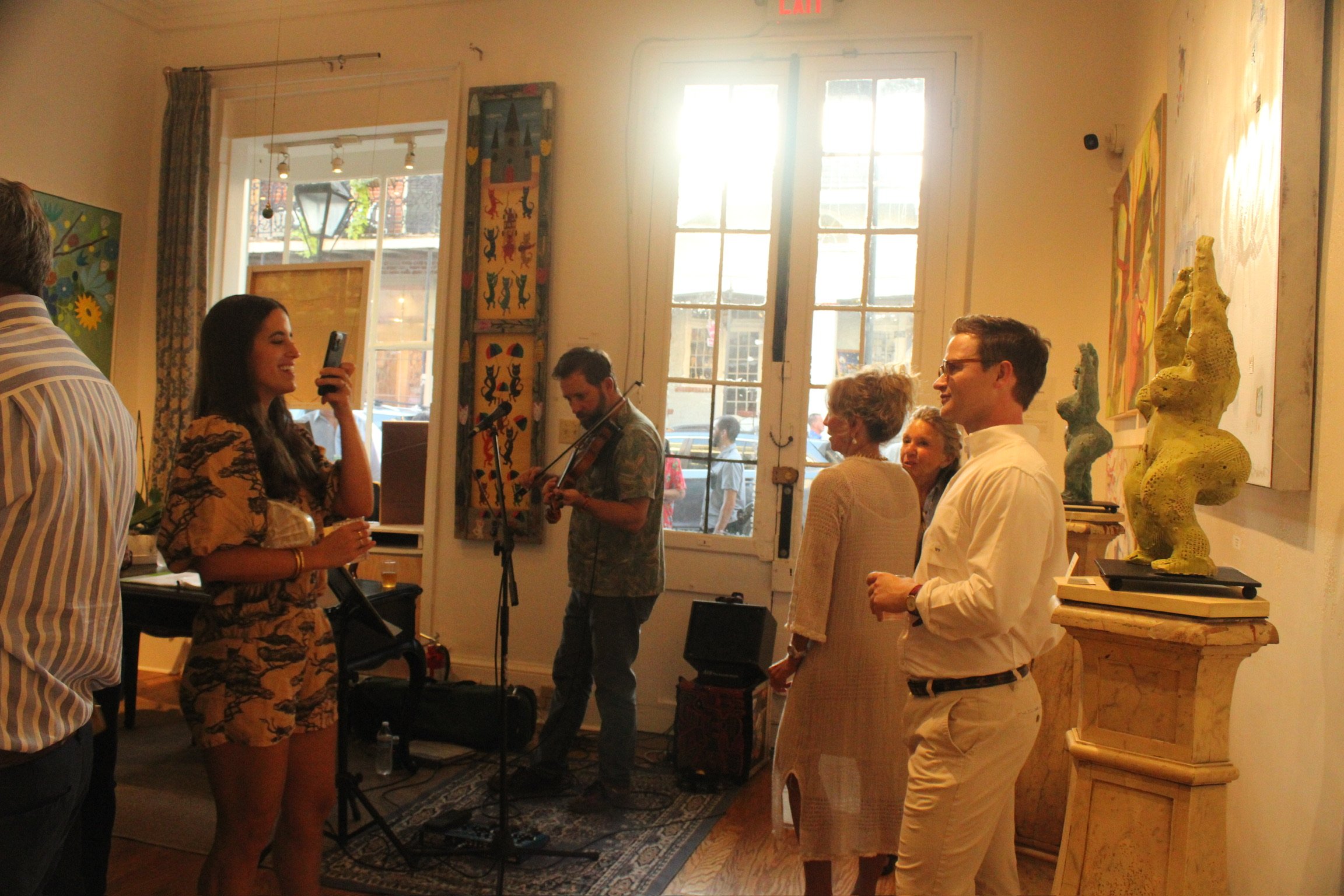 A group of people gathered in an art gallery, some engaging with each other and a musician playing a violin. The gallery has large windows allowing natural light, colorful artwork on the walls, and sculptures on display.