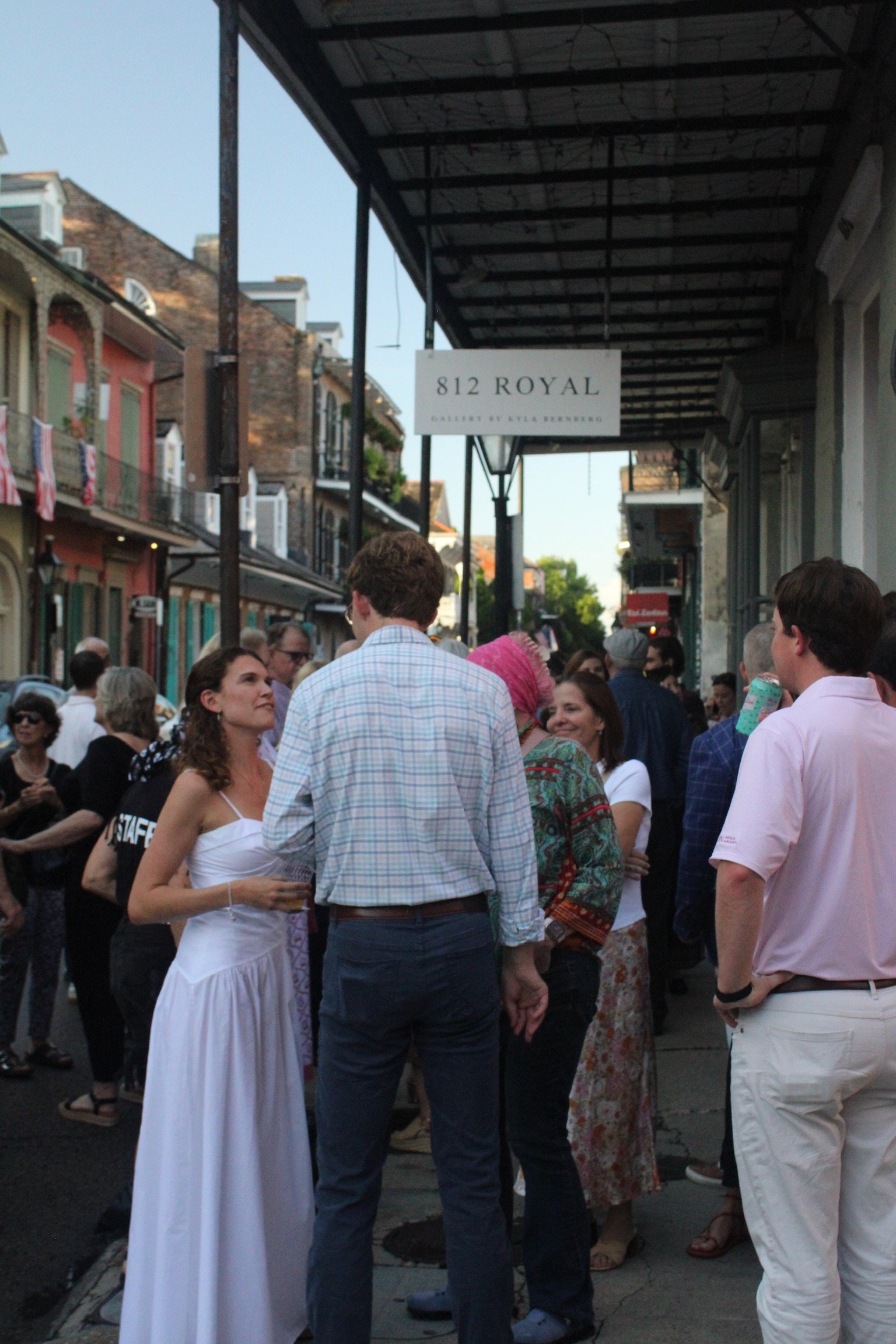People gathered on a street, some engaged in conversation, with buildings and a sign for '812 Royal Gallery by Eva Barnes' overhead.