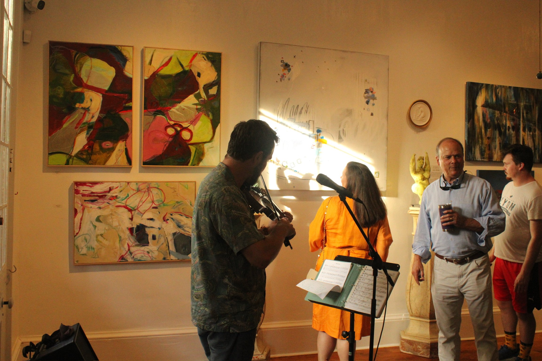 People at an art gallery with abstract paintings on the wall, some individuals talking and playing musical instruments, sunlight streaming through a window.