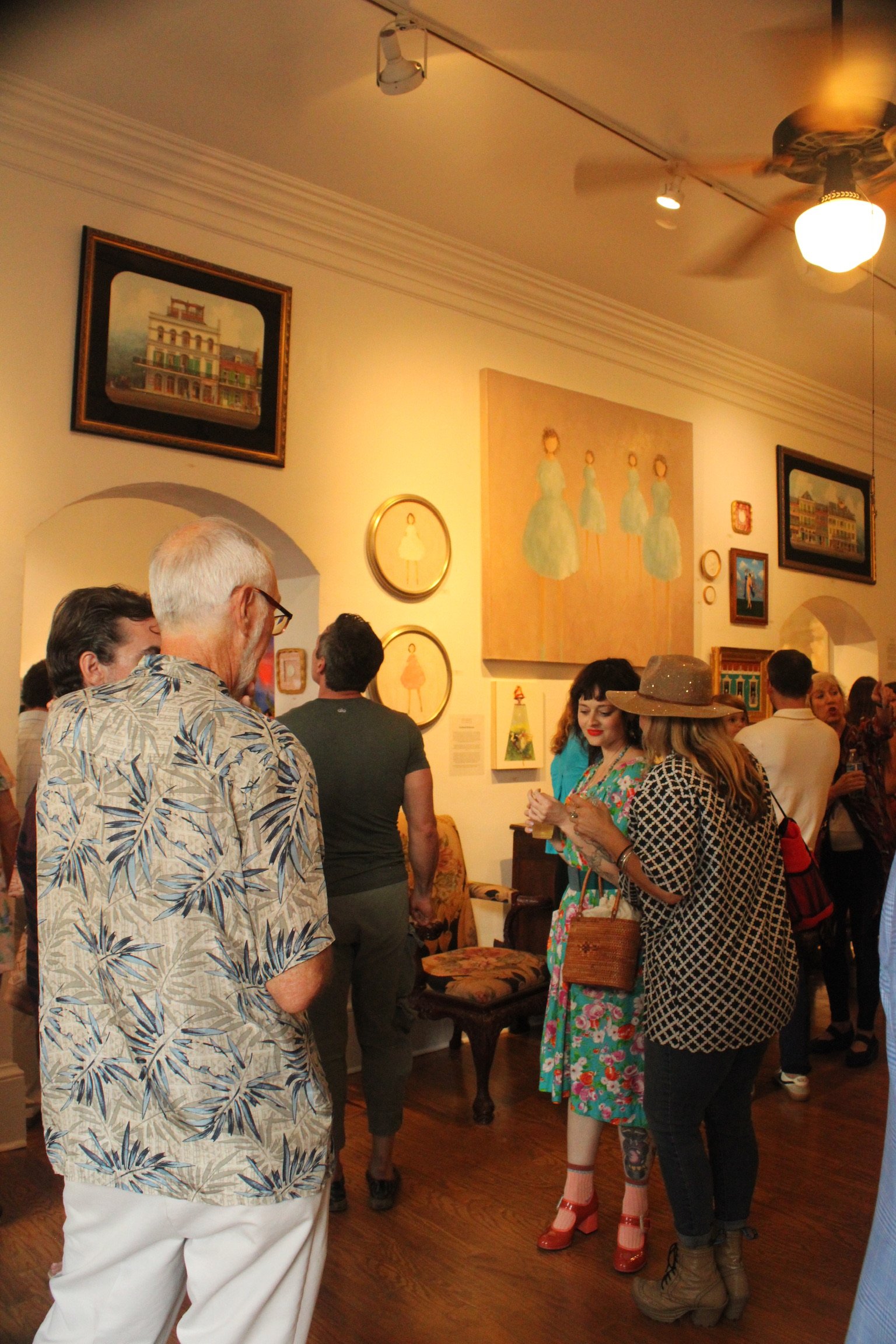 People gathered in an art gallery or museum, looking at paintings on the wall, with some engaged in conversation and wearing colorful and patterned clothing.