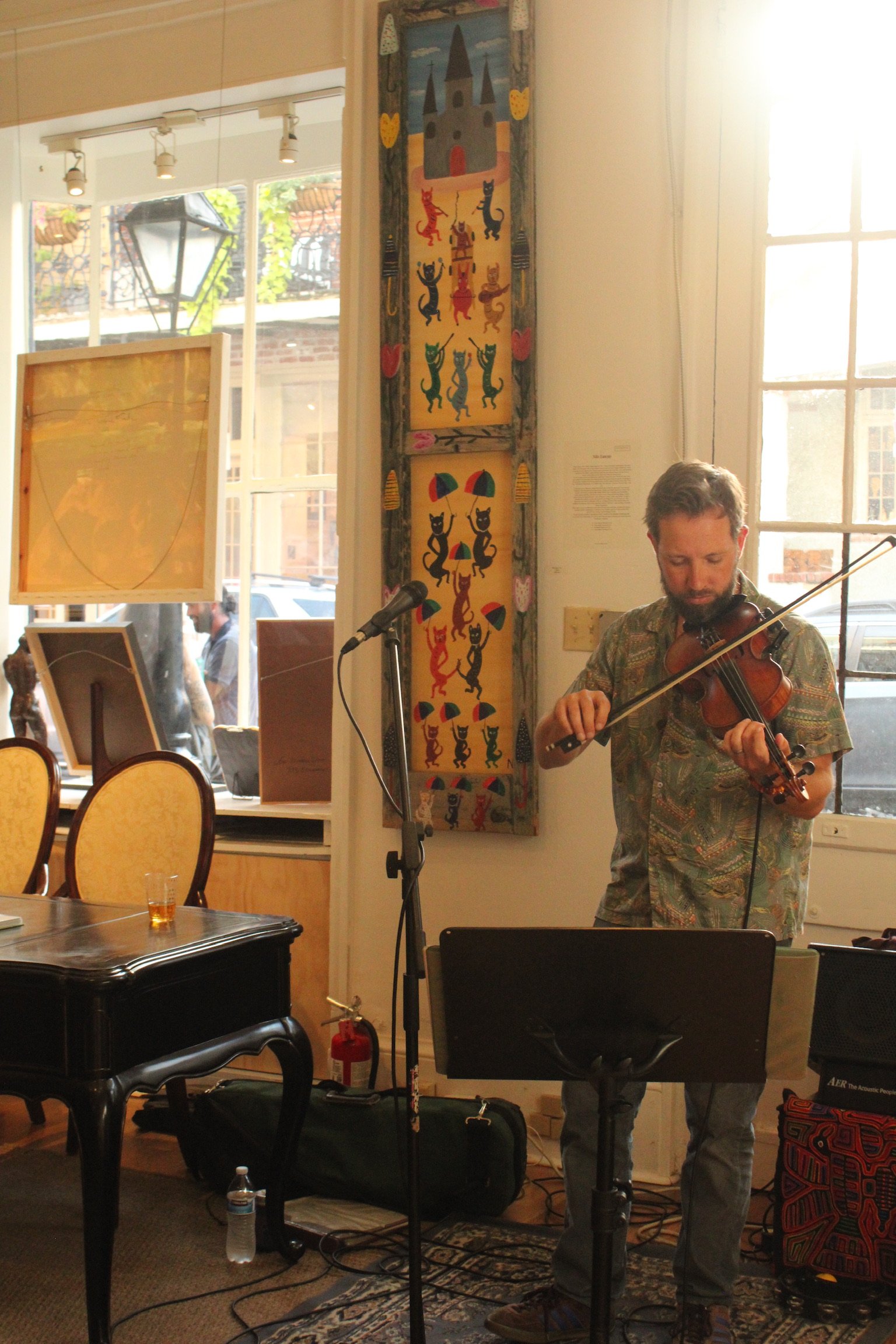 A man playing the violin at a small indoor event or performance, with musical stands, microphones, and colorful wall art featuring cats and a castle in the background.