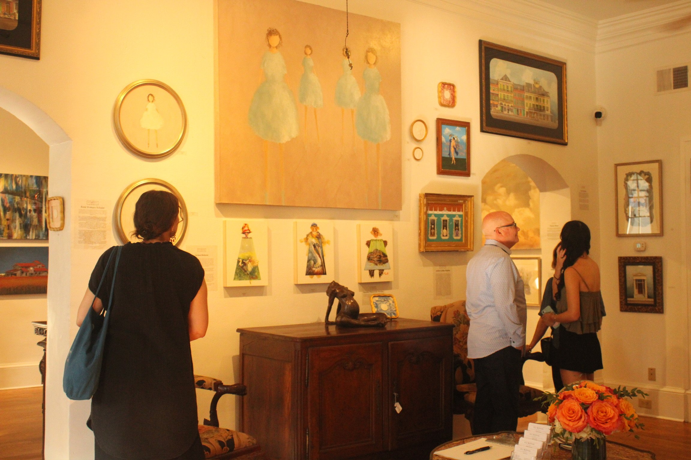 People viewing art in an art gallery with various paintings and framed artwork on the walls. A woman with black hair and a blue bag stands facing the art, while an older man and a woman with dark hair speak near a table with a bouquet of orange roses