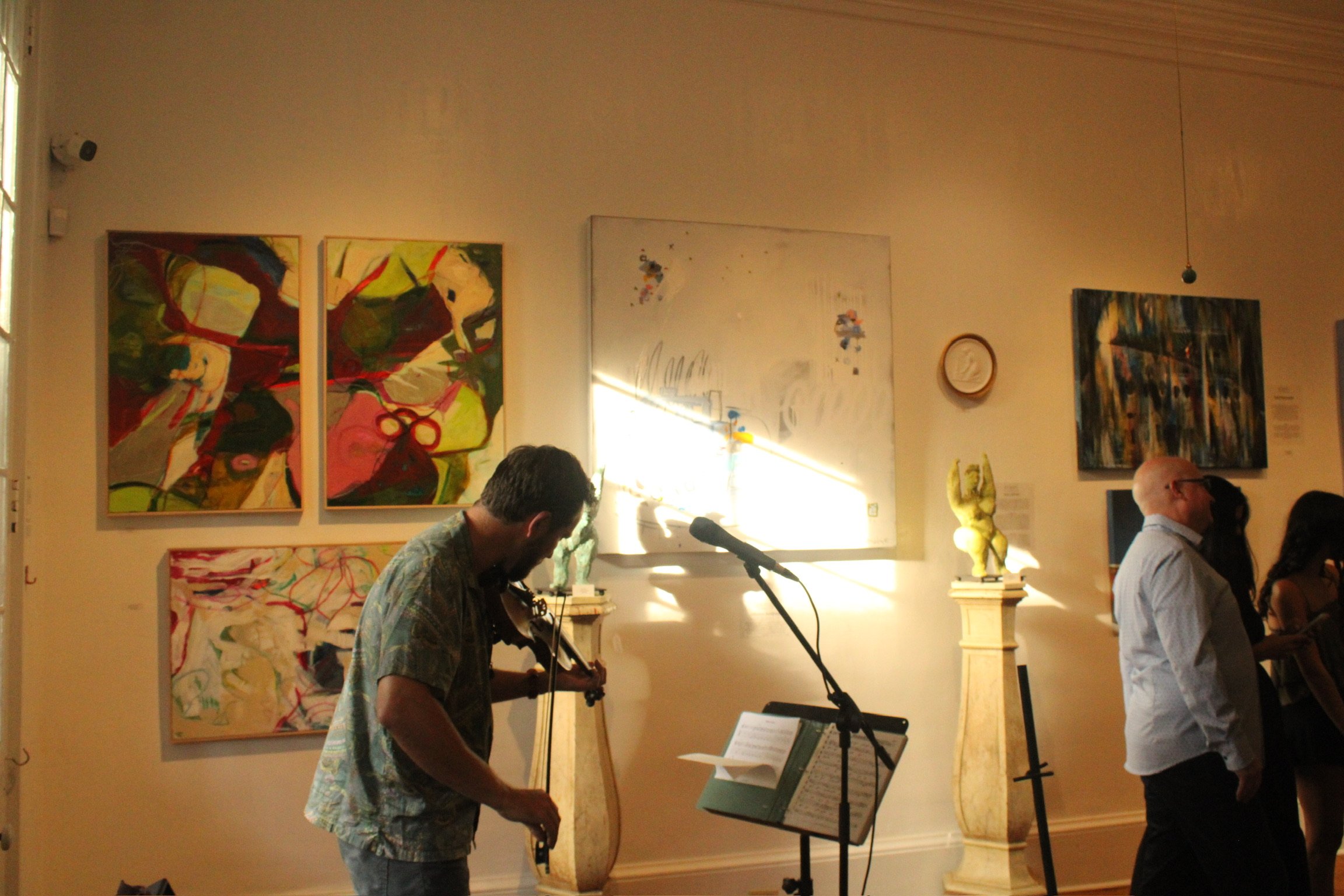 A musical performance at an art gallery with paintings on the wall. A man is playing the guitar, a woman is playing the violin, and people are standing and watching.