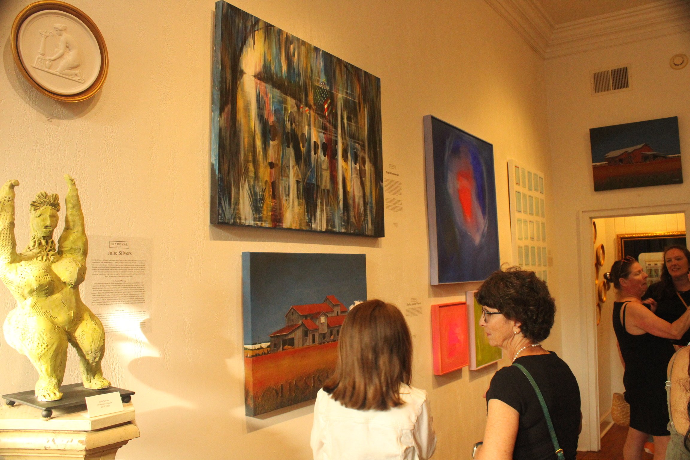 People viewing artwork in an art gallery, including a large abstract painting, a traditional landscape, and a red barn scene, with sculptures and gallery information plaques visible.