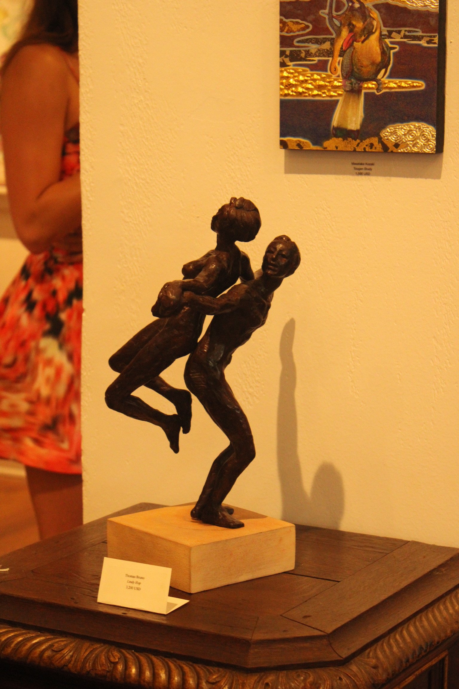 Bronze sculpture of two figures in a dance pose, situated on a wooden base on a table. In the background, part of a wall with artwork and a person in a floral dress is visible.