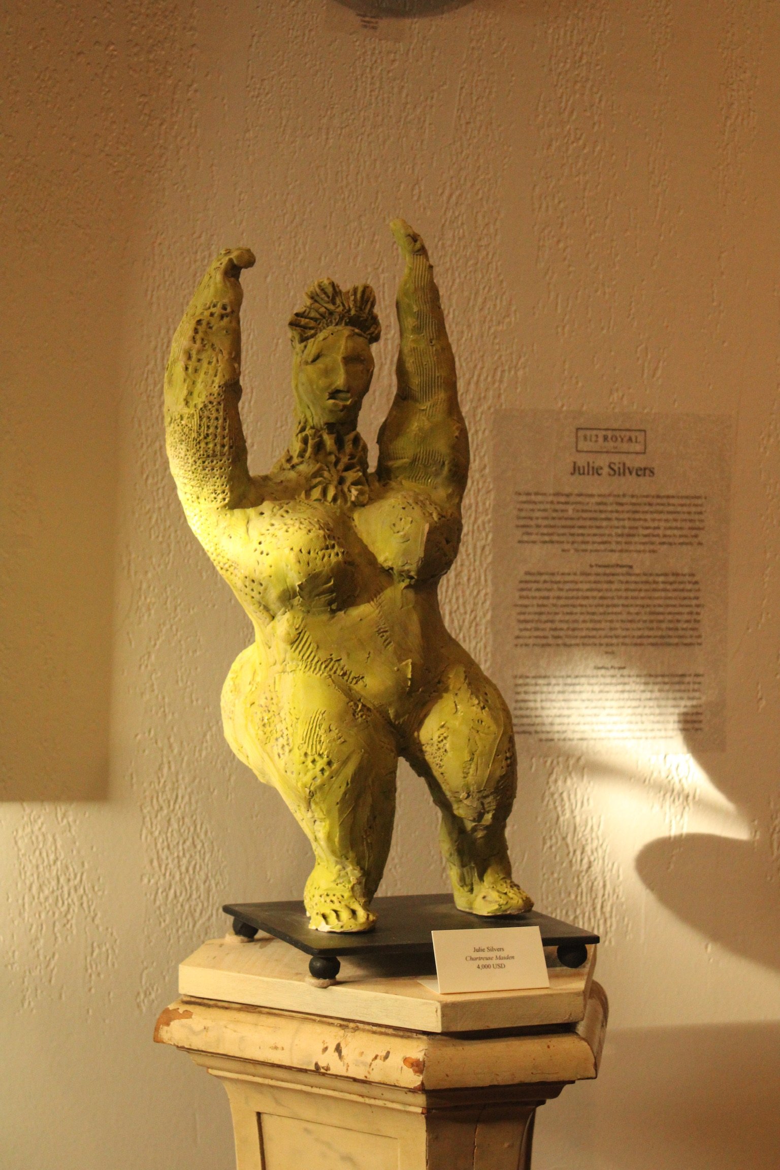 A sculpture titled 'Charismatic Manners' by Julie Silvers, priced at 4,000 USD, displayed on a pedestal in a gallery. The sculpture resembles a stylized figure with exaggerated features and textured, yellowish-green skin.