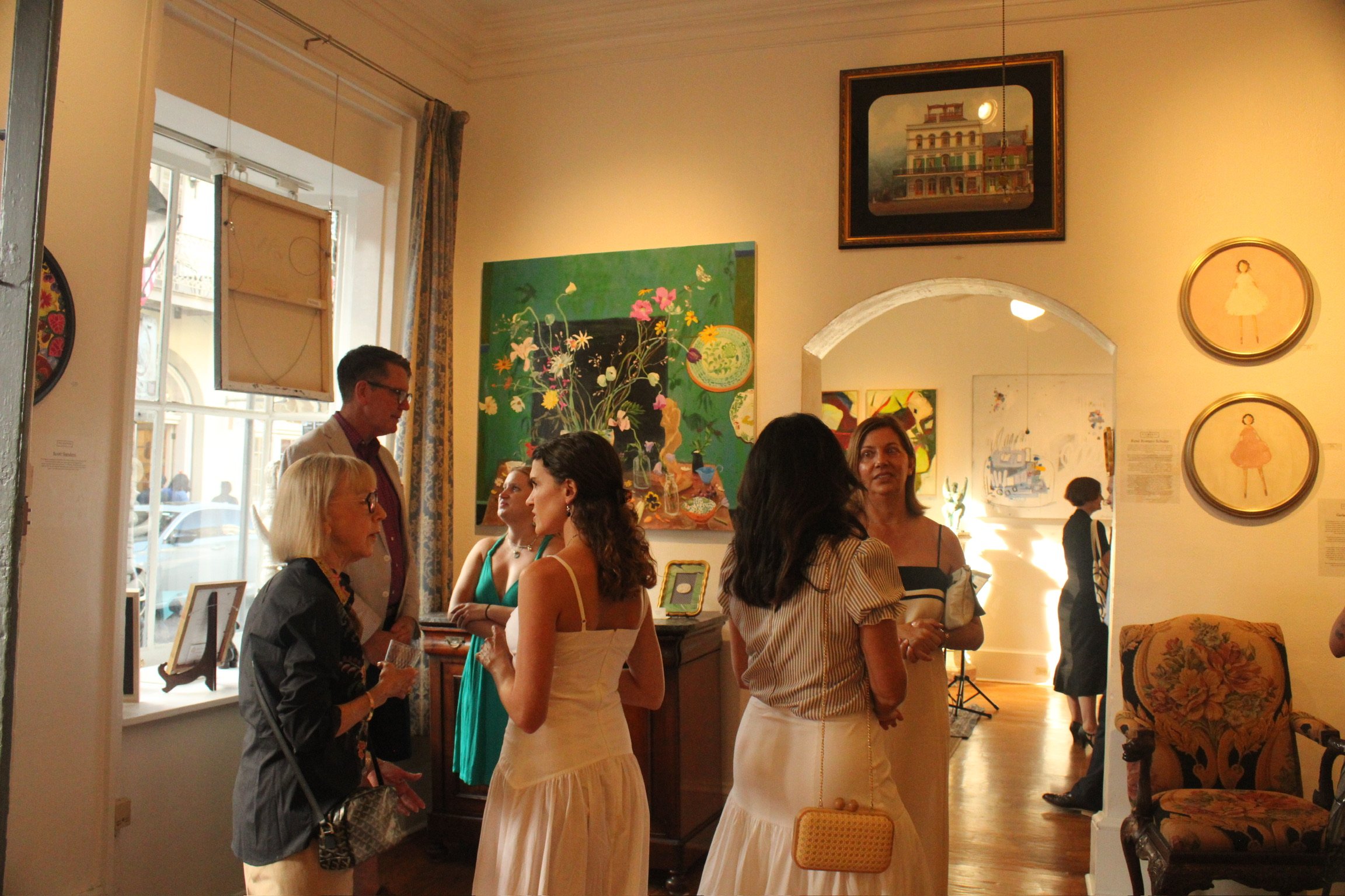 Group of people inside an art gallery engaging in conversation, with various paintings and framed artwork on the walls, and a large window letting in natural light.