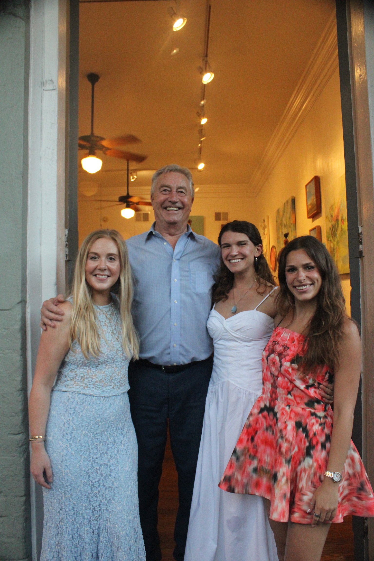 A group of five people, four women and one man, smiling and standing together indoors, with art on the walls behind them.