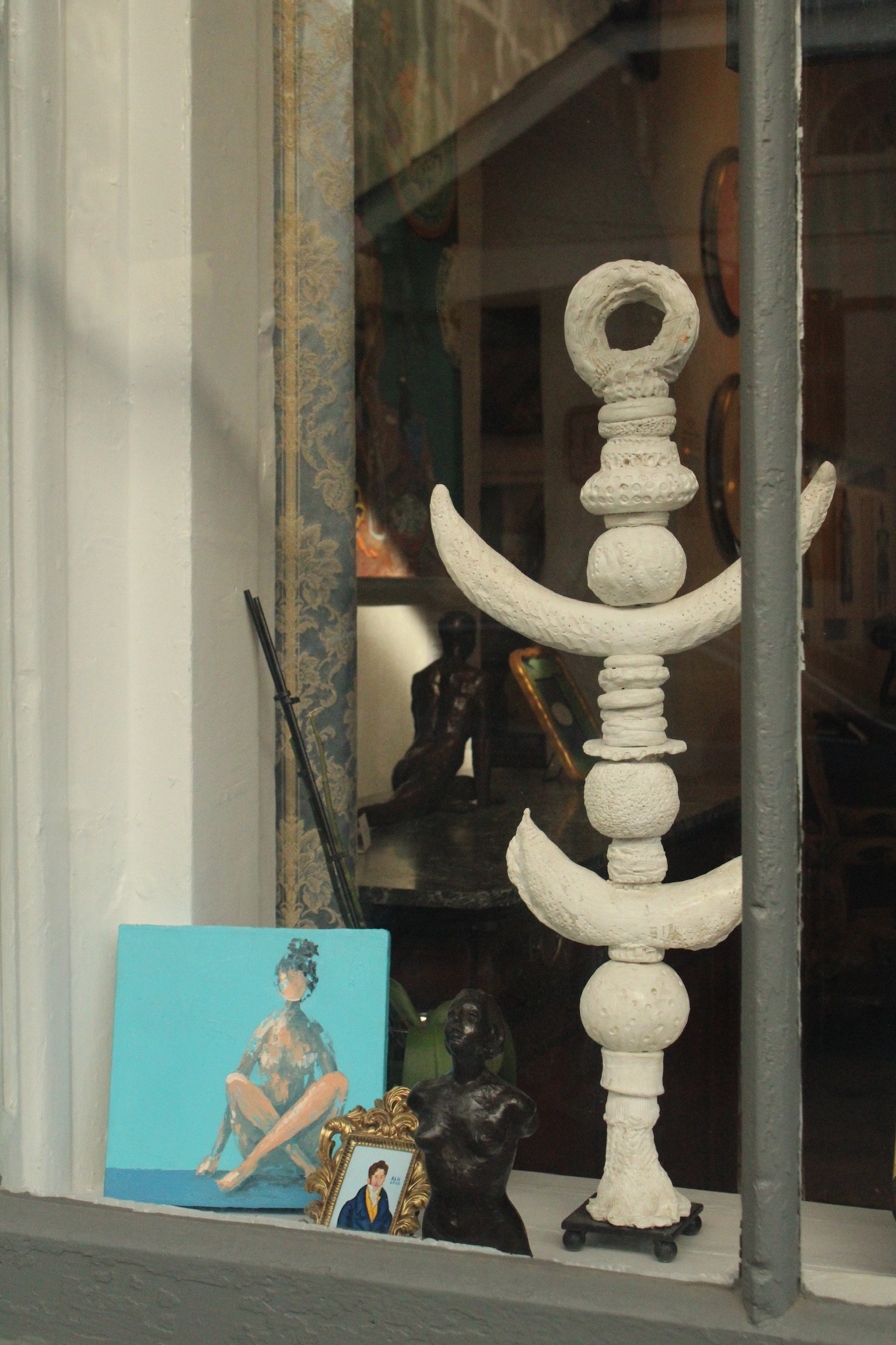 View of a storefront display containing various artworks and sculptures, including a tall white sculpture resembling an anchor, a painting of a person sitting, and framed portraits, visible through a window with metal bars.