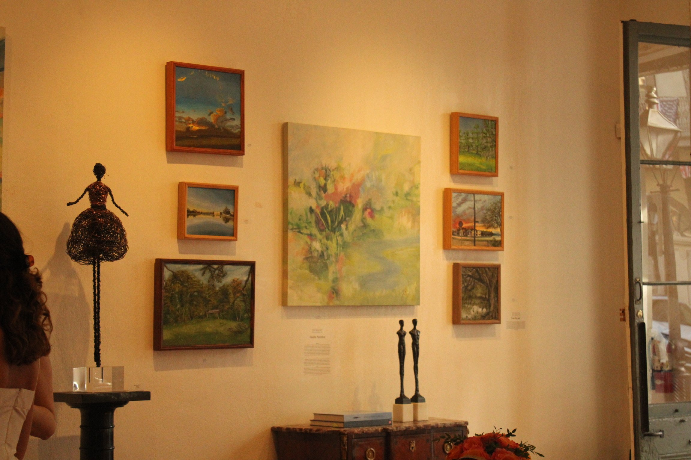 An art gallery wall displaying various landscape paintings in frames, with a sculpture of a woman on a stand and two tall candlesticks on a table, seen through a glass door in a warmly lit room.