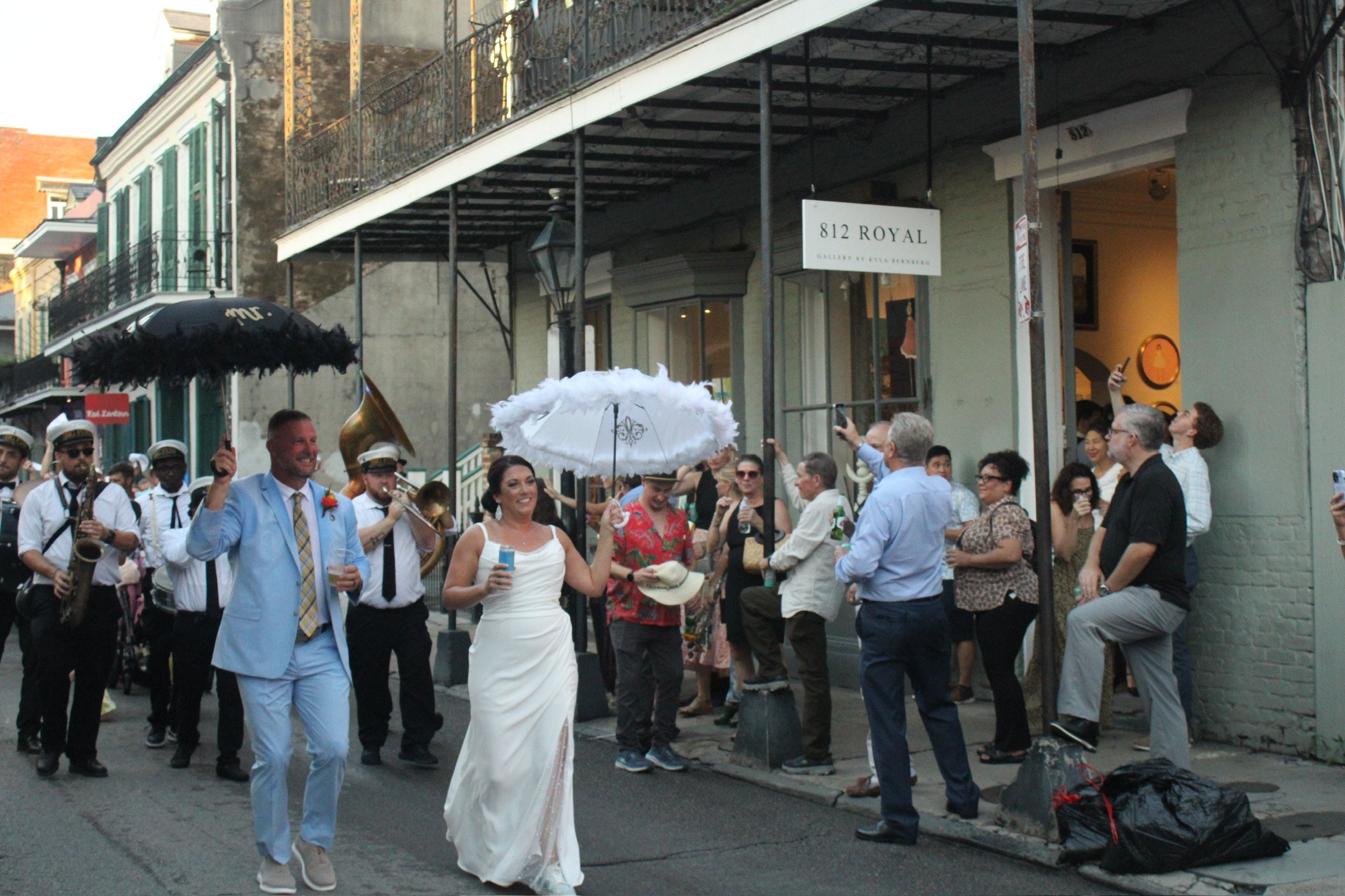 Street scene with people dressed in wedding attire, a band playing, and a crowd gathering for a celebration on a city sidewalk in front of a gallery at 812 Royal.