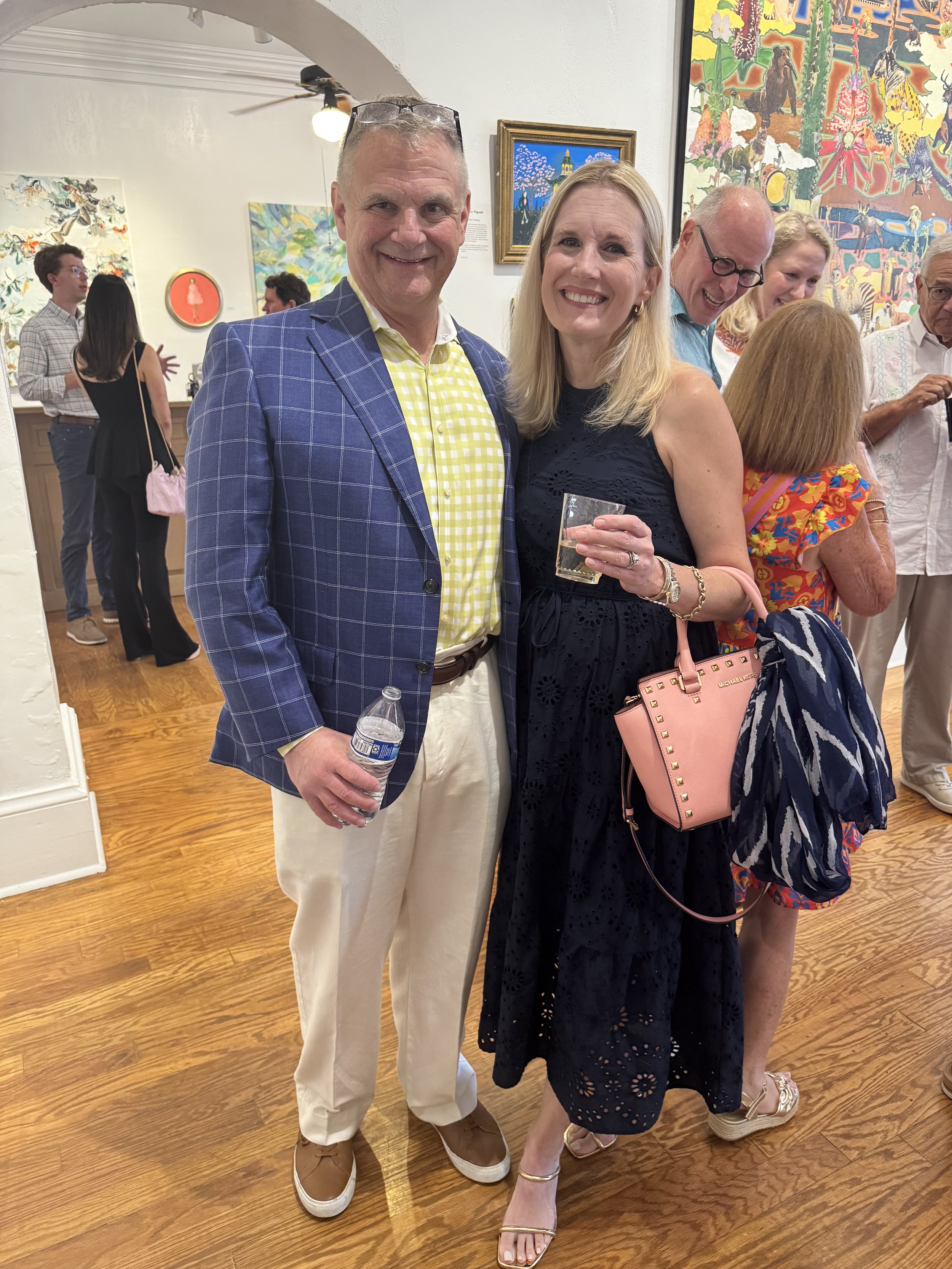 A group of people gathered in an art gallery, with two people in the foreground smiling at the camera. The man is wearing a blue checkered blazer, yellow checkered shirt, and beige pants, holding a water bottle. The woman is wearing a black dress, go