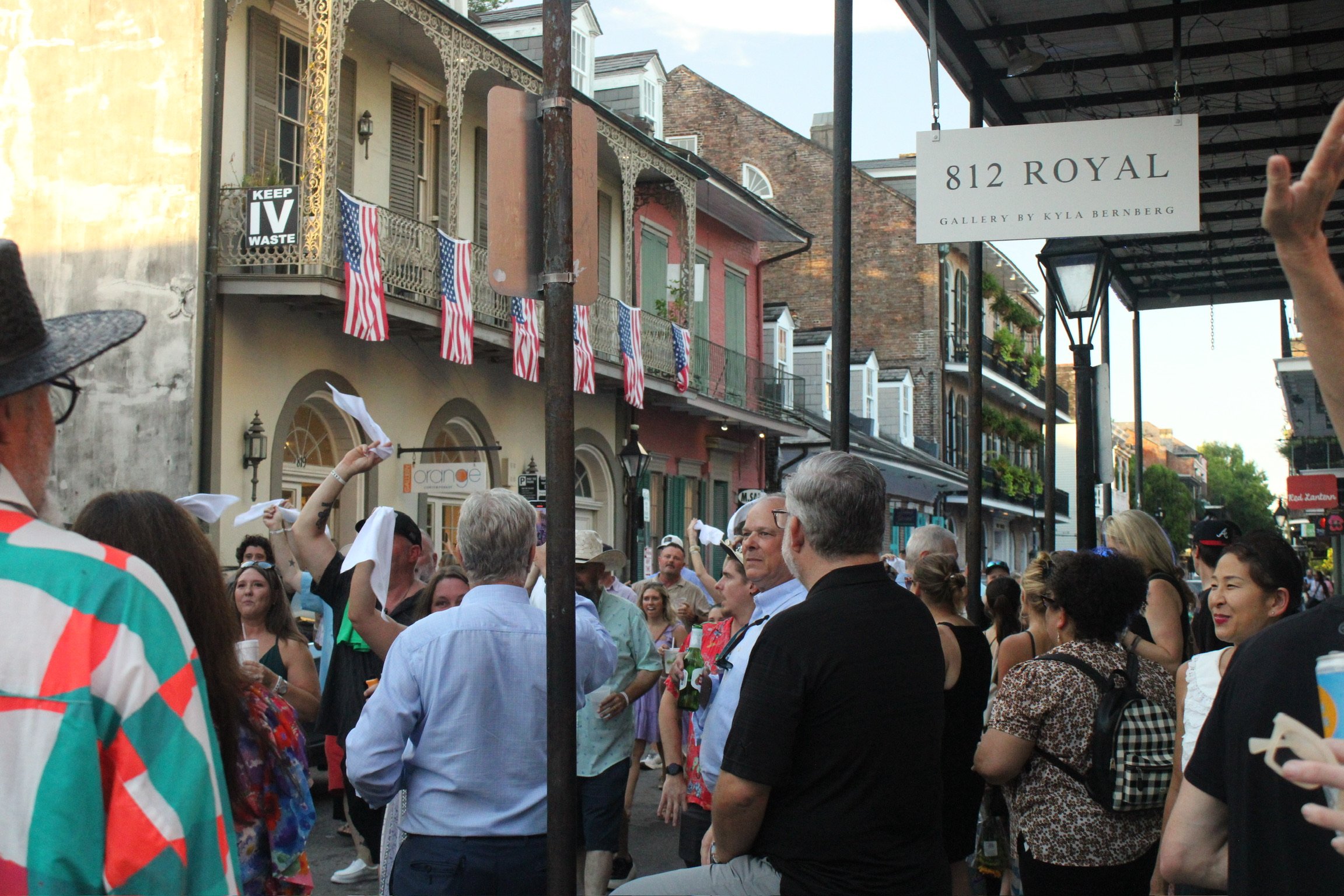 Crowd of people gathered outside on a city street, some holding flags, surrounded by historic buildings with balconies decorated with American flags, and a sign that reads '812 Royal Gallery by Kyla Bernberg'.