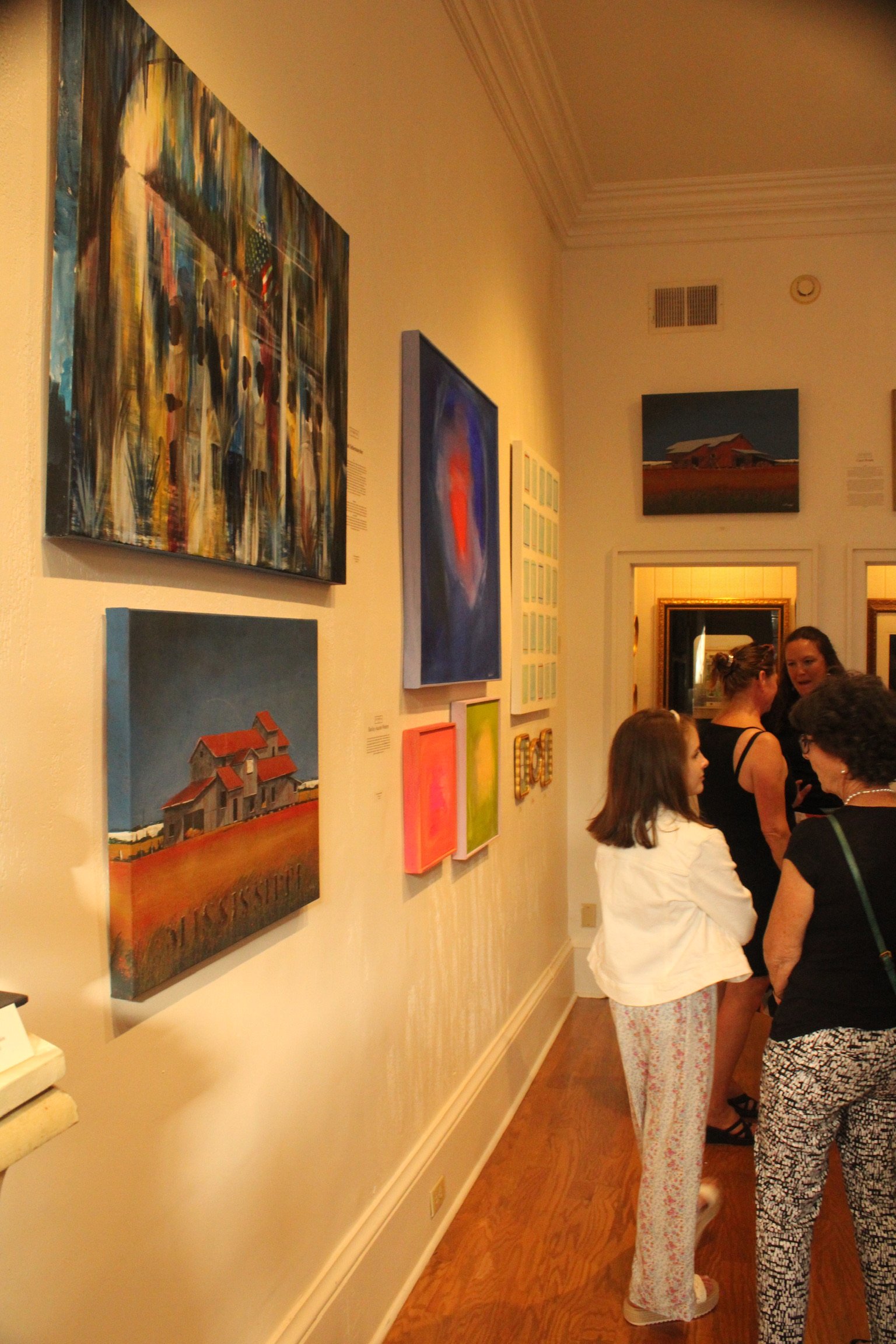 People viewing artwork in a gallery with paintings on the walls, including a colorful abstract piece and a landscape with a barn.