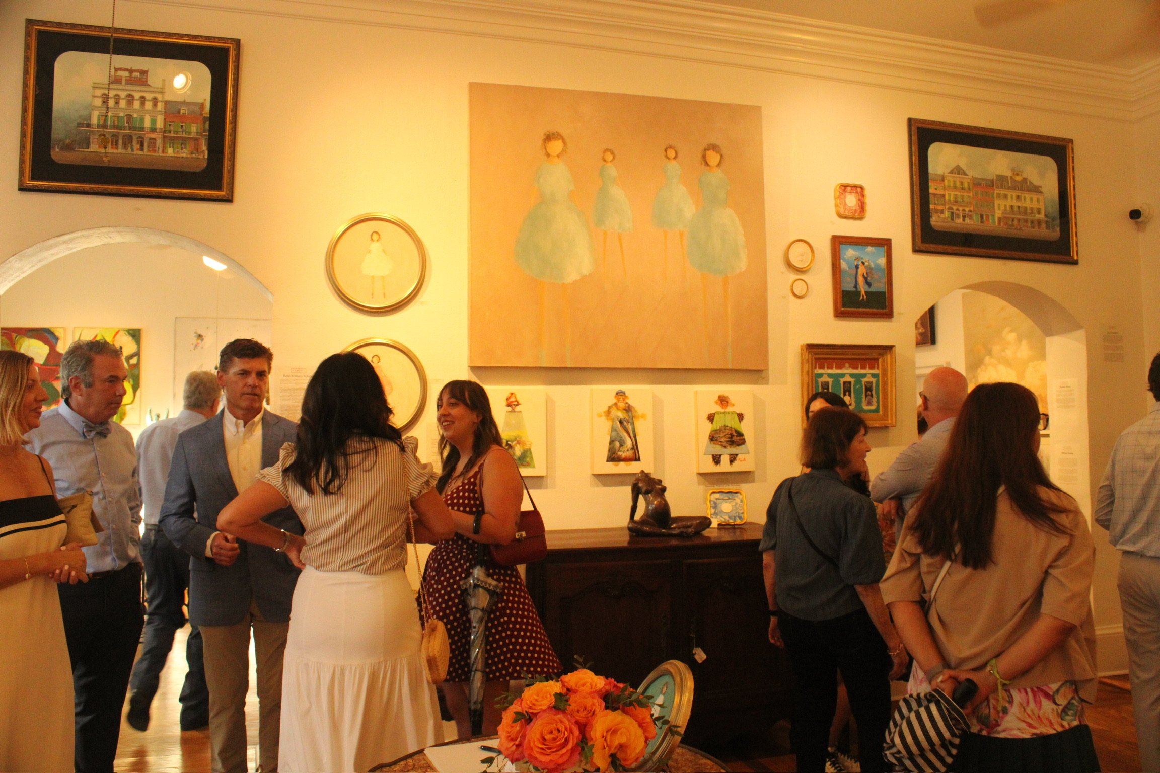 People attending an art gallery, engaging in conversation, with various paintings on the walls, a large floral arrangement on a table, and a warmly lit interior.