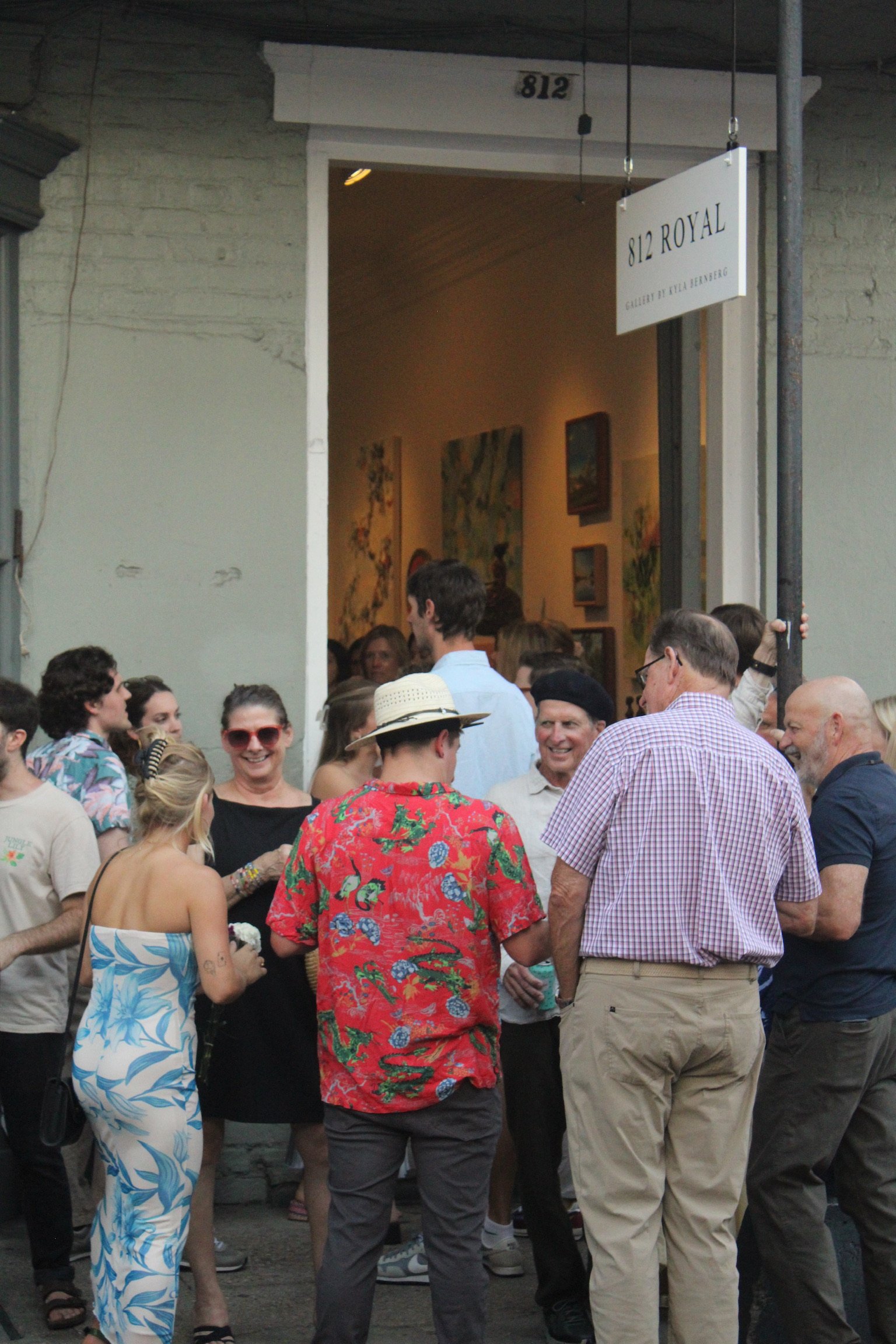 People gathered outdoors at an art gallery opening, with some engaging in conversation, and artworks hanging on the interior wall visible through the door.