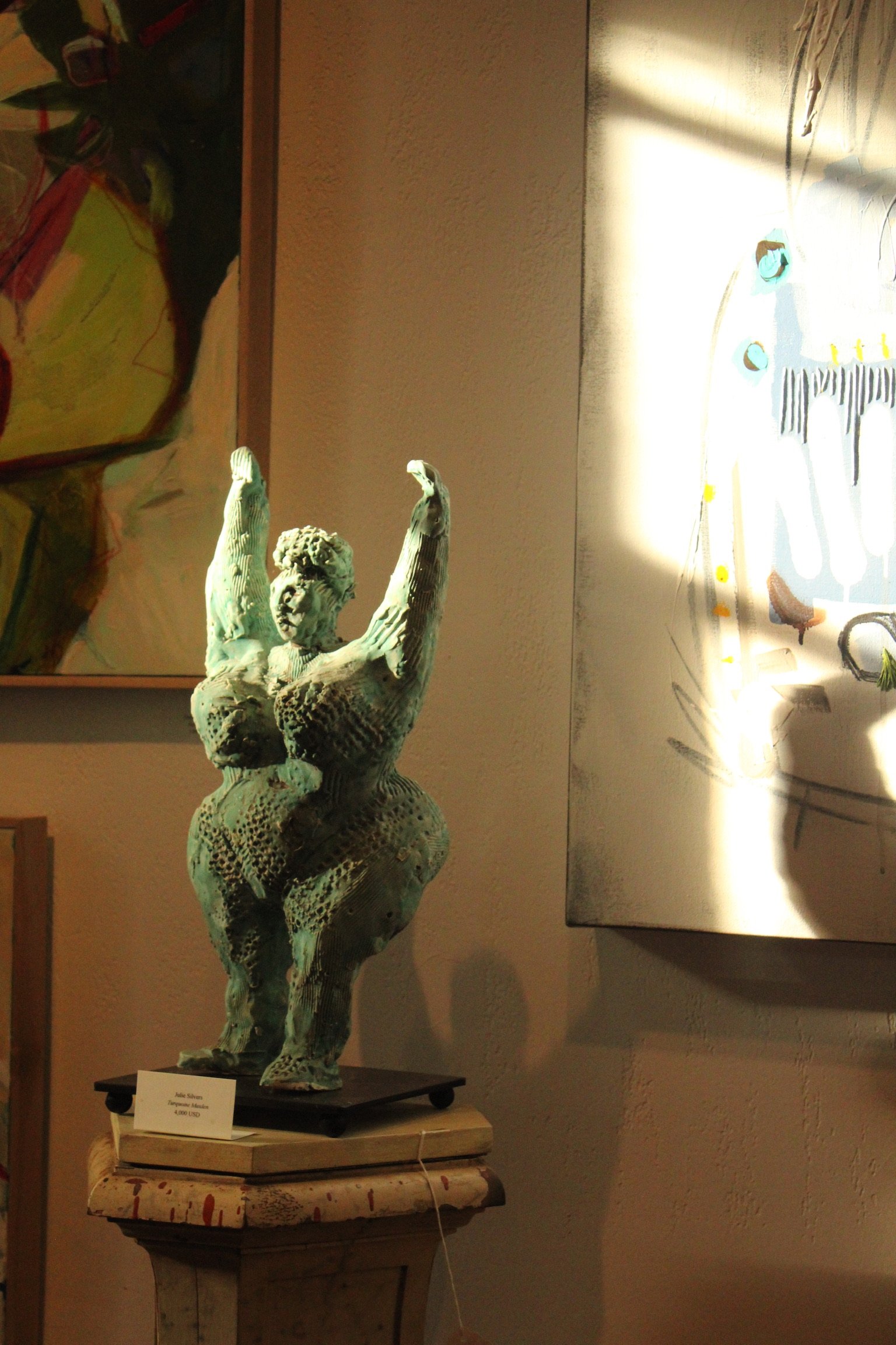 A ceramic sculpture of a woman with arms raised, placed on a pedestal. Artwork is displayed behind, with a light illuminating one side of the piece.