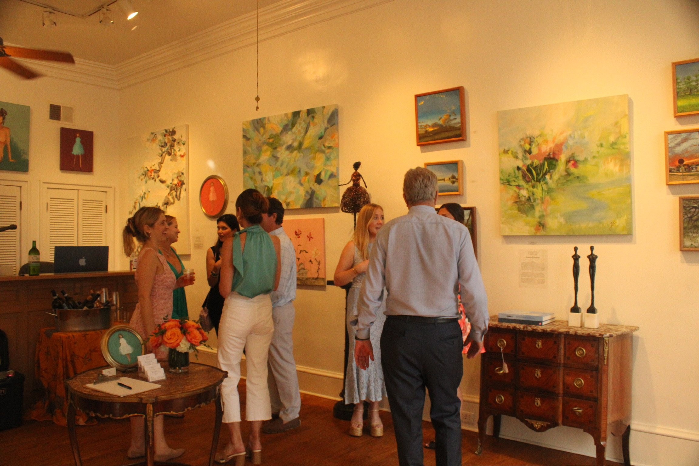 People attending an art gallery exhibition, looking at colorful paintings on the walls, with some individuals engaged in conversation.