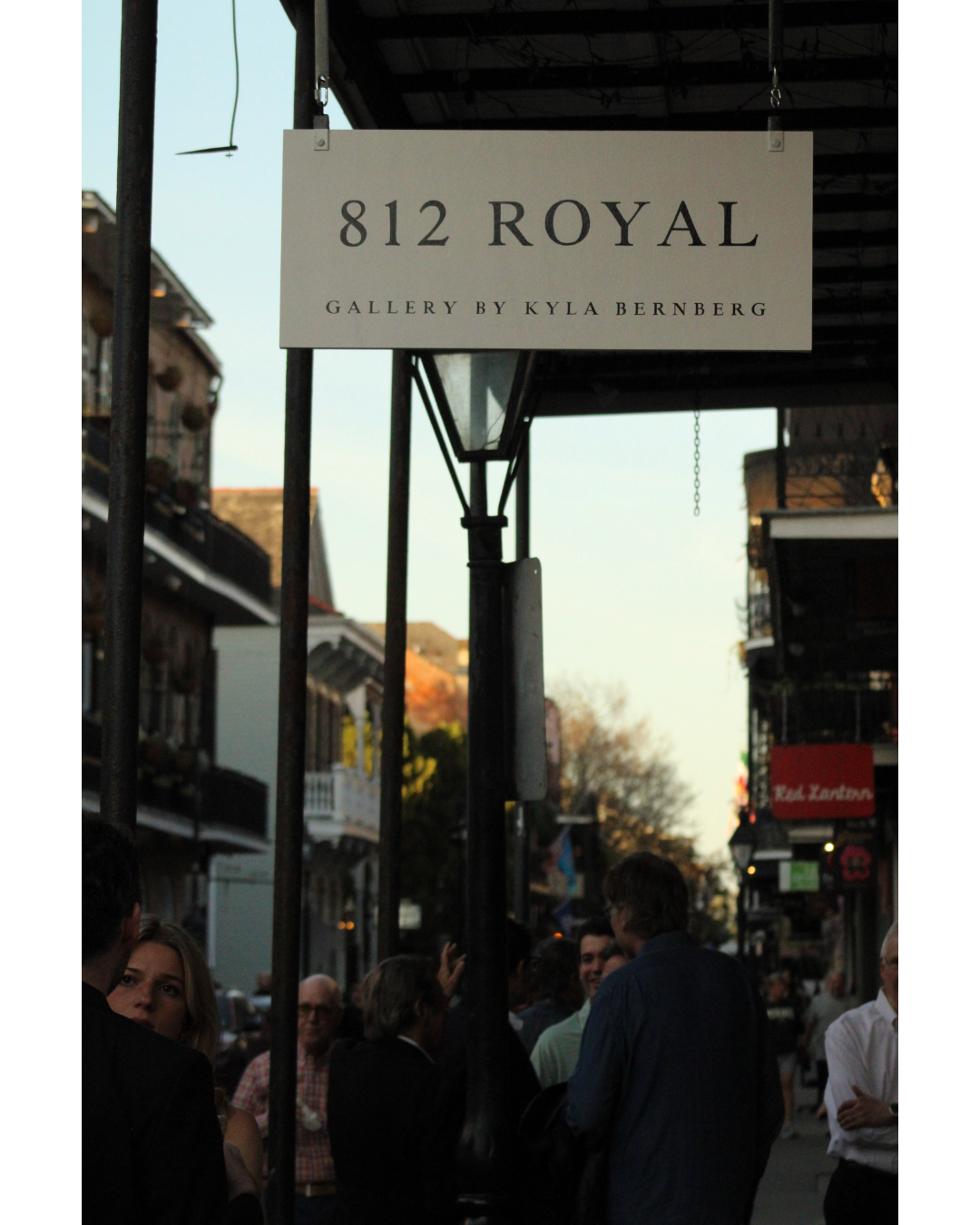 A street scene with people gathering under a sign that reads '812 Royal, Gallery by Kyla Bernberg' on a city street with shops and apartments.