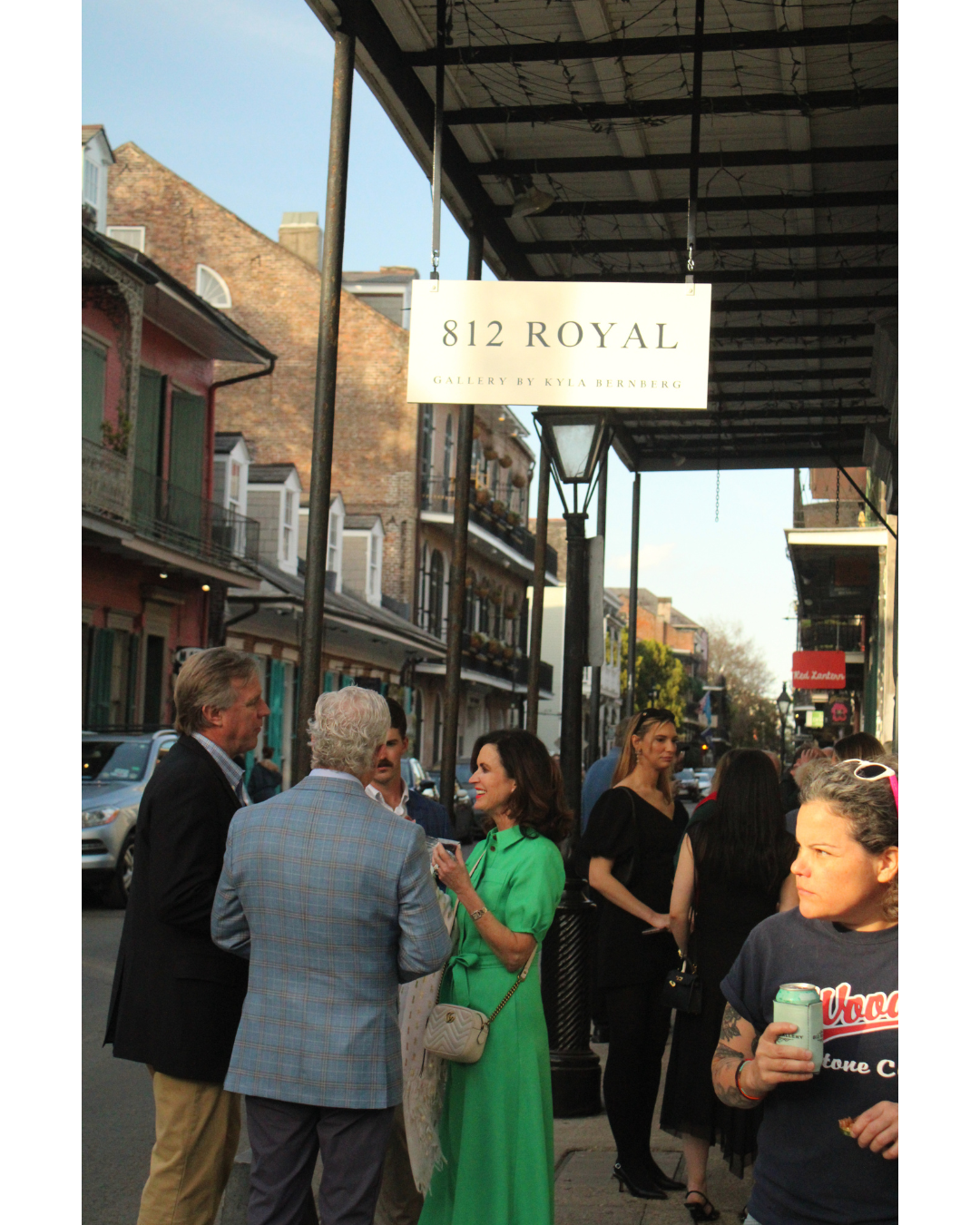 People standing on a sidewalk in front of a building with a sign that reads '812 Royal Gallery by Kyla Bernstein' in an urban setting with shops and balconies.