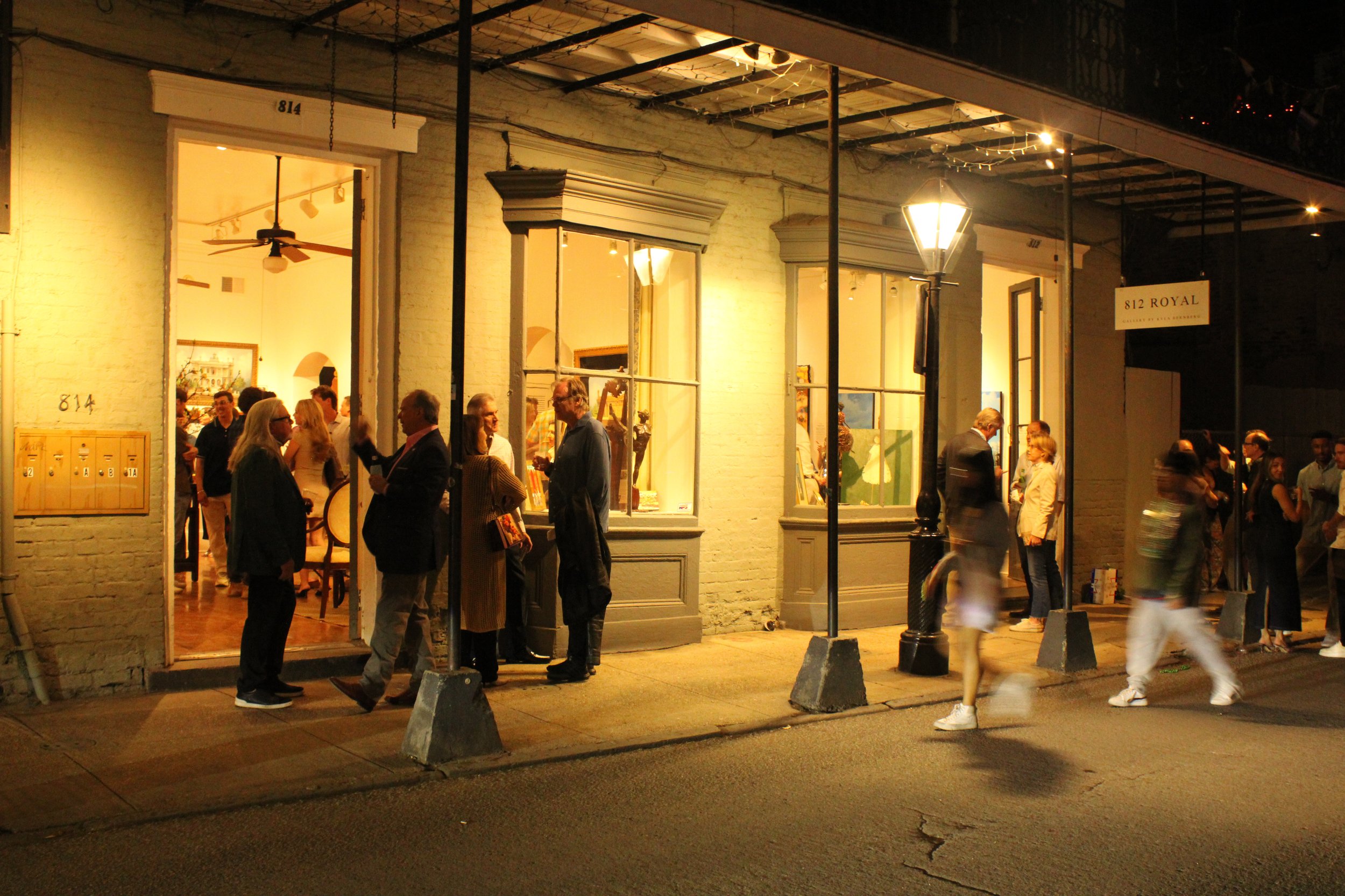 People gather outside a brightly lit art gallery or shop at night on a city street, with some people in conversation and others walking by.