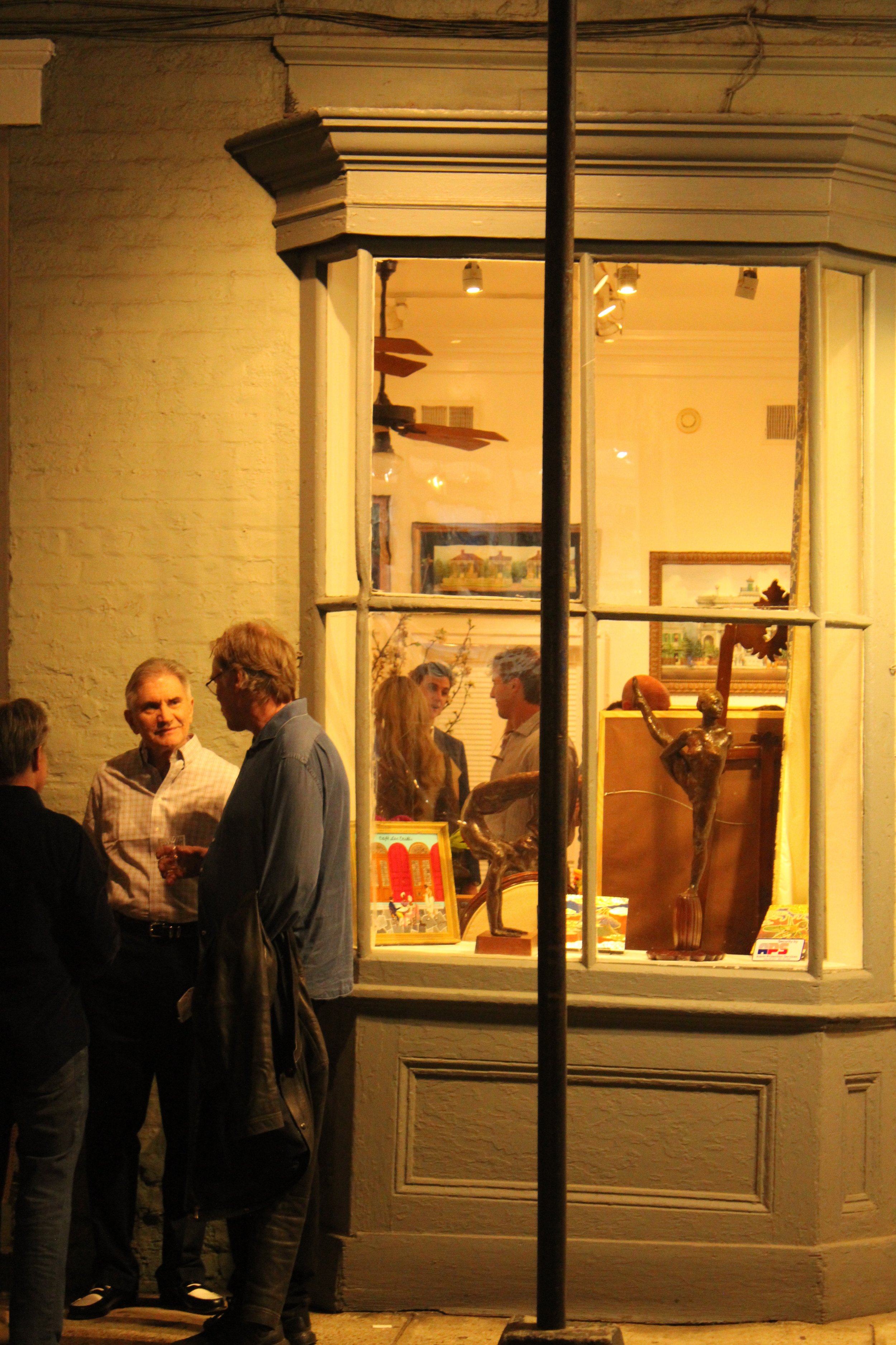 People standing outside and talking in front of a store window at night, with various sculptures and art pieces visible inside.