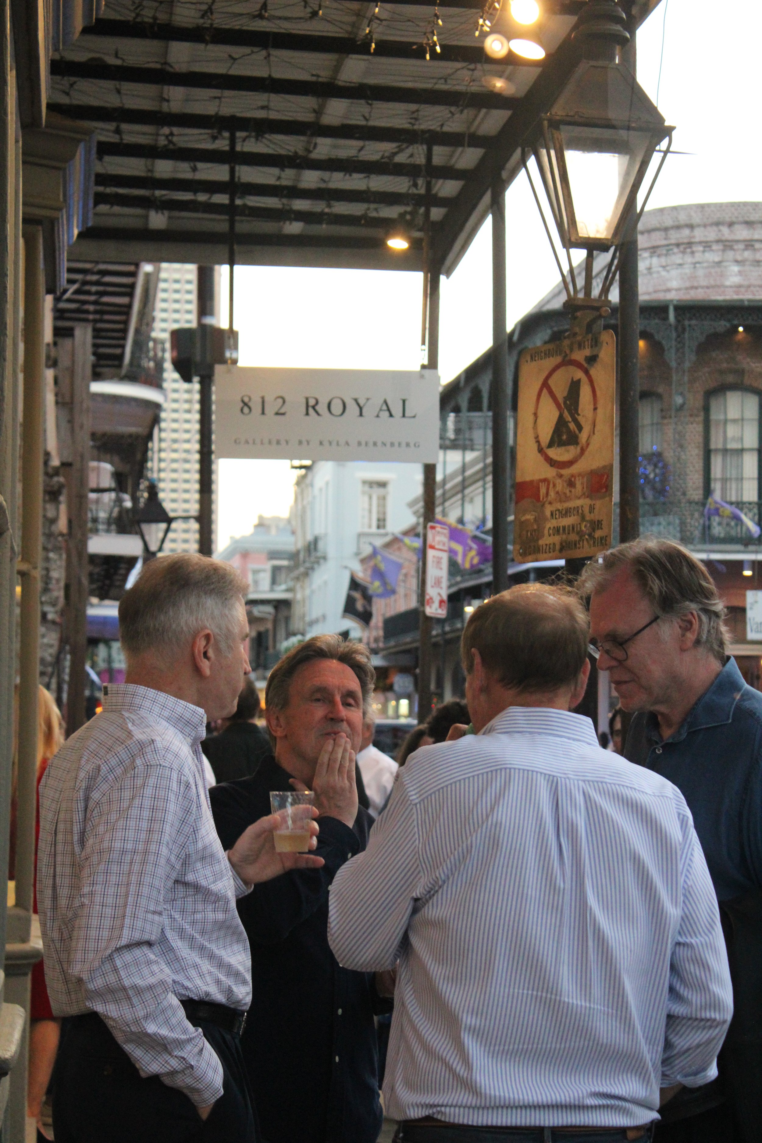 A group of men standing together outdoors at a street event, in conversation, with some holding drinks. The background shows a sign reading '812 Royal,' and other street signs and buildings, indicating an urban setting.