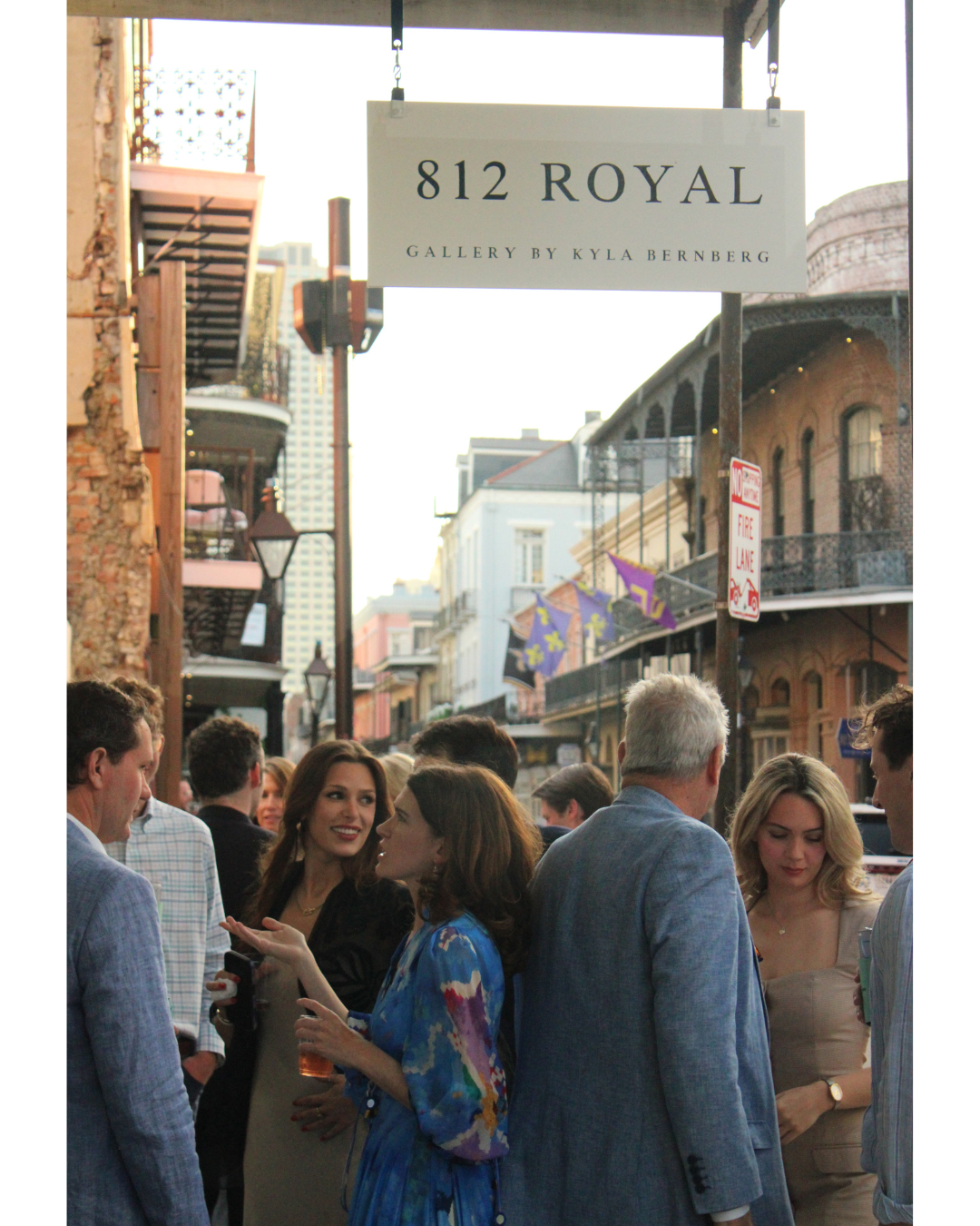 A group of people gathering on a busy street in front of a gallery called "812 Royal" with a sign overhead, during daylight hours. The street is lined with historic buildings and decorative flags.
