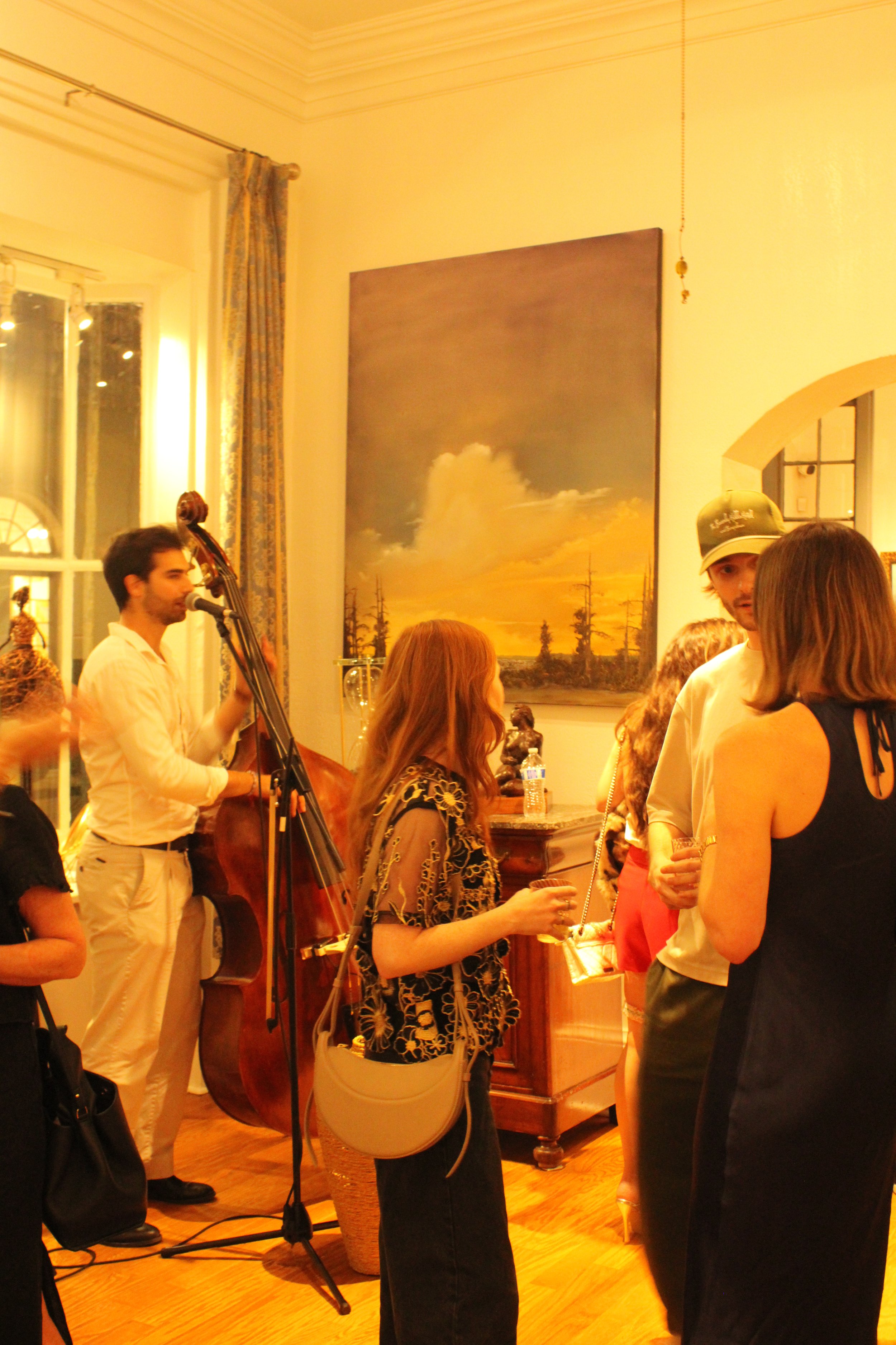 A group of people socializing at an indoor gathering with a live band playing. The band includes a man playing a double bass and singing into a microphone. The room has wooden floors, large windows, and a large landscape painting on the wall.