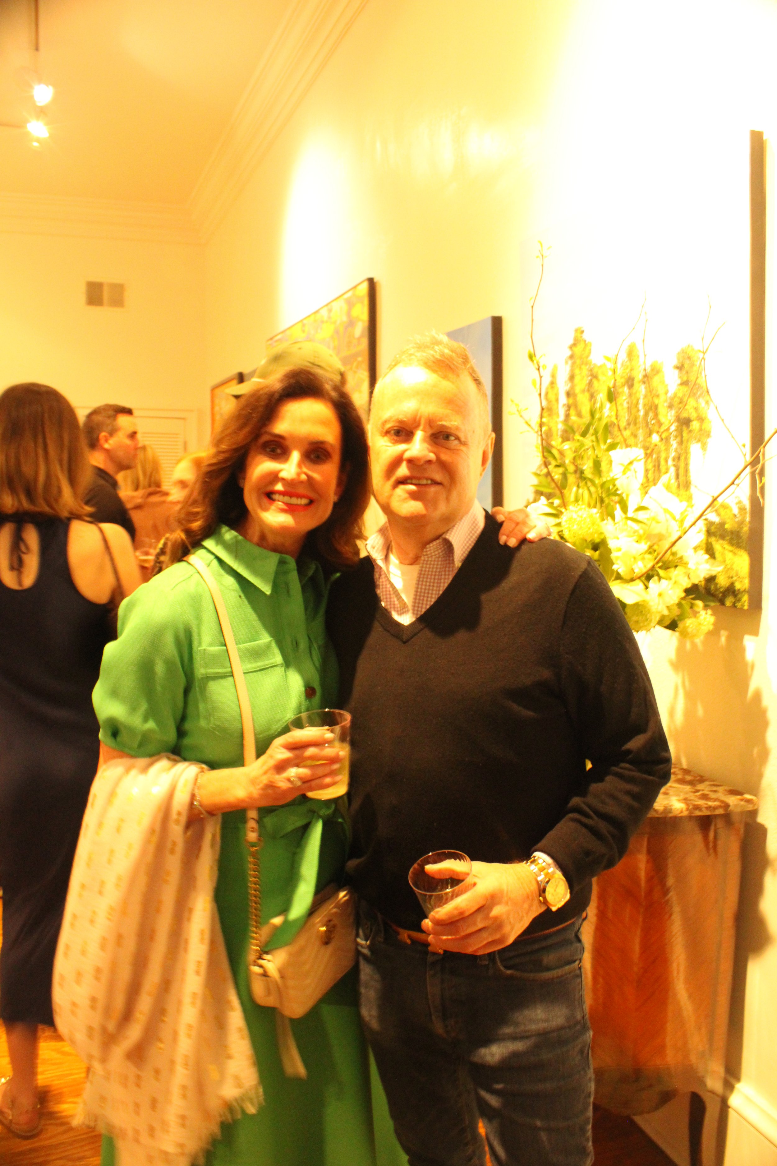 A woman in a bright green dress and a man in a dark sweater and jeans are smiling and holding drinks at a social gathering inside a well-lit room with yellow walls and floral artwork.