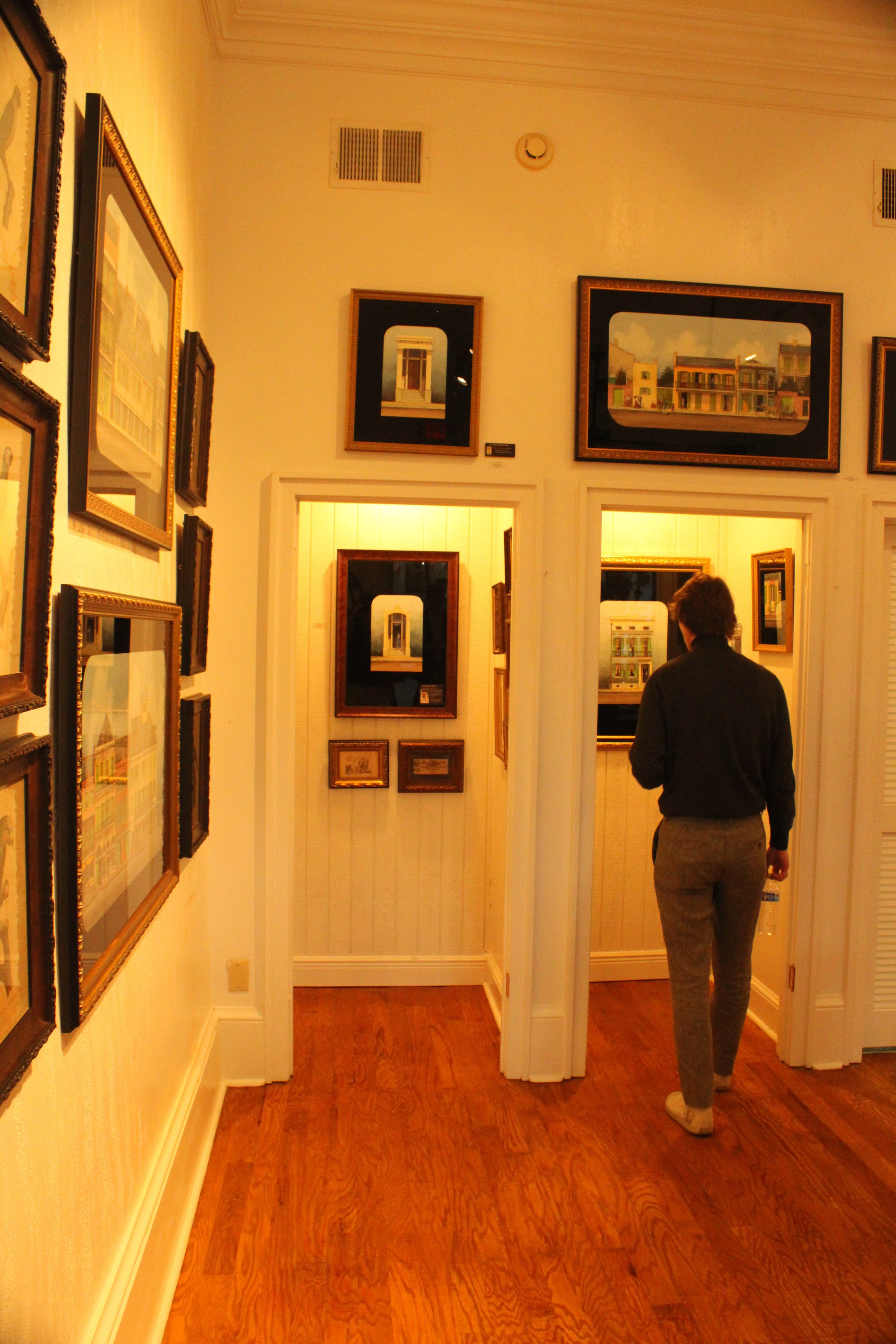 A person is standing in an art gallery, looking at framed artwork on a yellow wall. The gallery has wooden floors and a high ceiling.