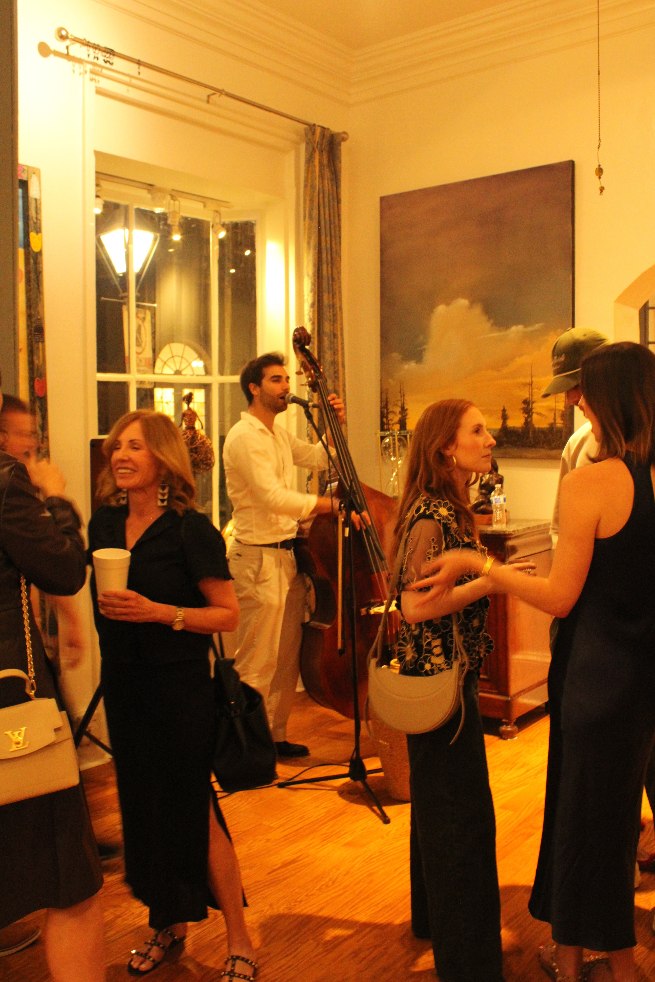 Group of people socializing at a gathering, with a musician playing a double bass in the background, inside a warmly lit room with large window and painting on wall.