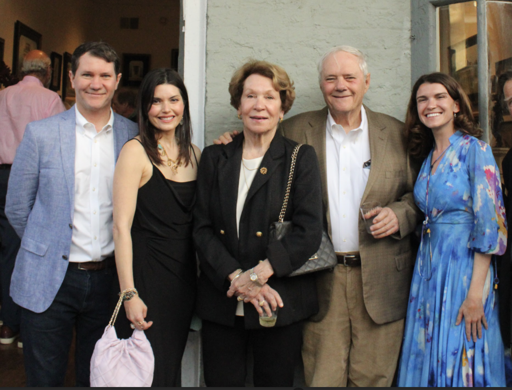 Group of six people smiling and posing for a photo at an indoor event, standing against a wall. They are dressed in semi-formal attire.