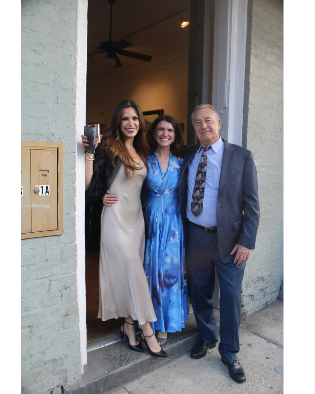 Three people, two women and one man, standing together at the entrance of a building, smiling at the camera. The woman on the left is holding a glass and has long, wavy brown hair, wearing a beige dress with black high heels. The woman in the middle 