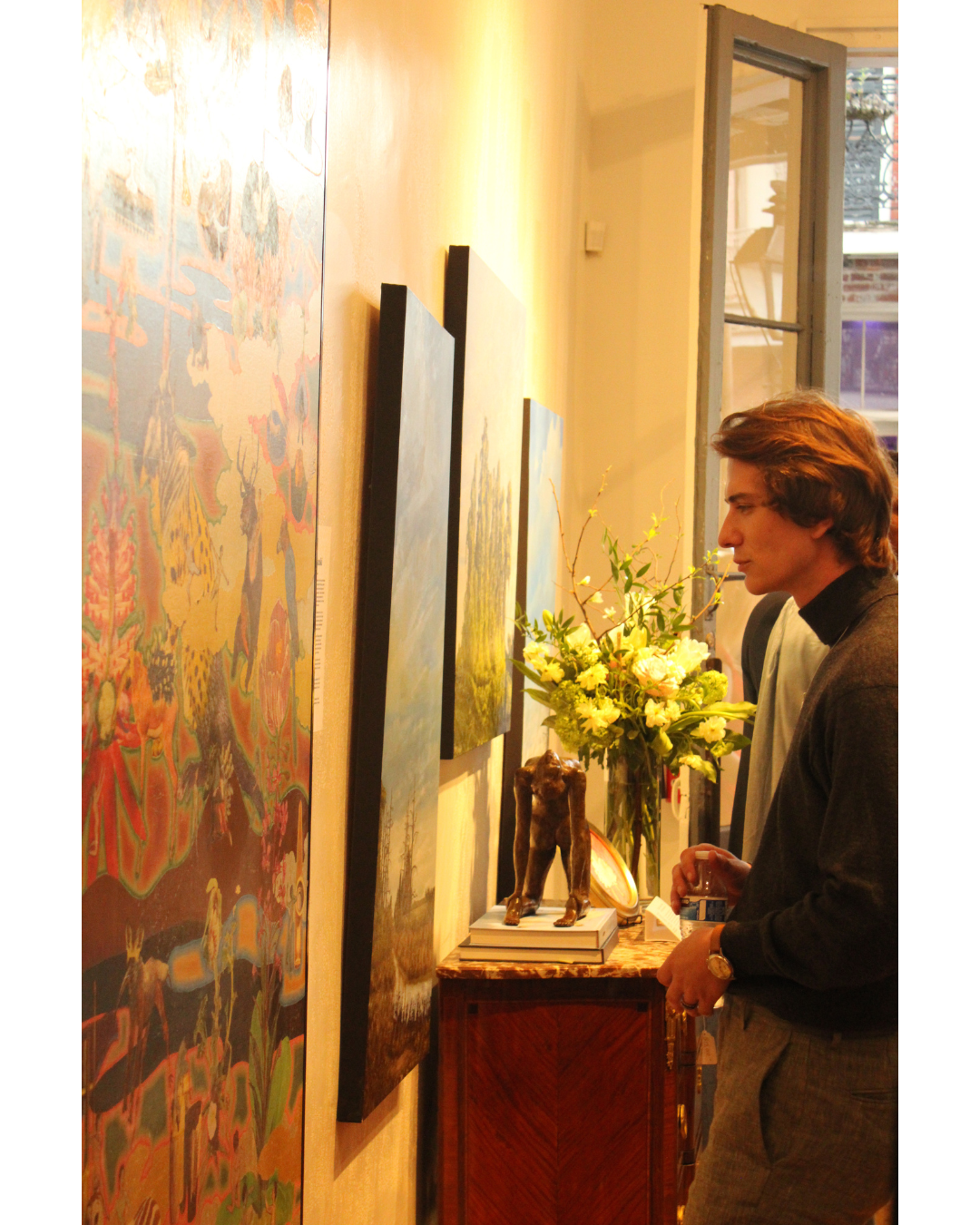 A man with brown hair, wearing a dark blazer, looking at artwork in an art gallery. He is holding a smaller item in his hand, standing near a wooden table with a flower arrangement, a sculpture, and stacked books.