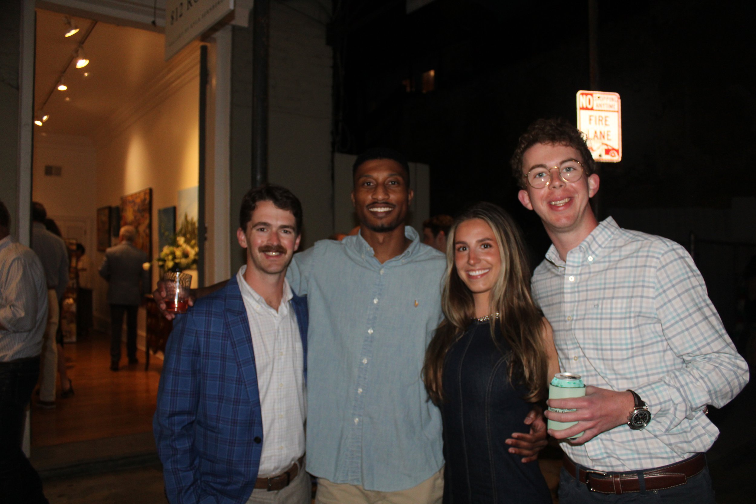 Group of four young adults smiling and posing together at night, one holding a drink in a sleeve, at an art opening.