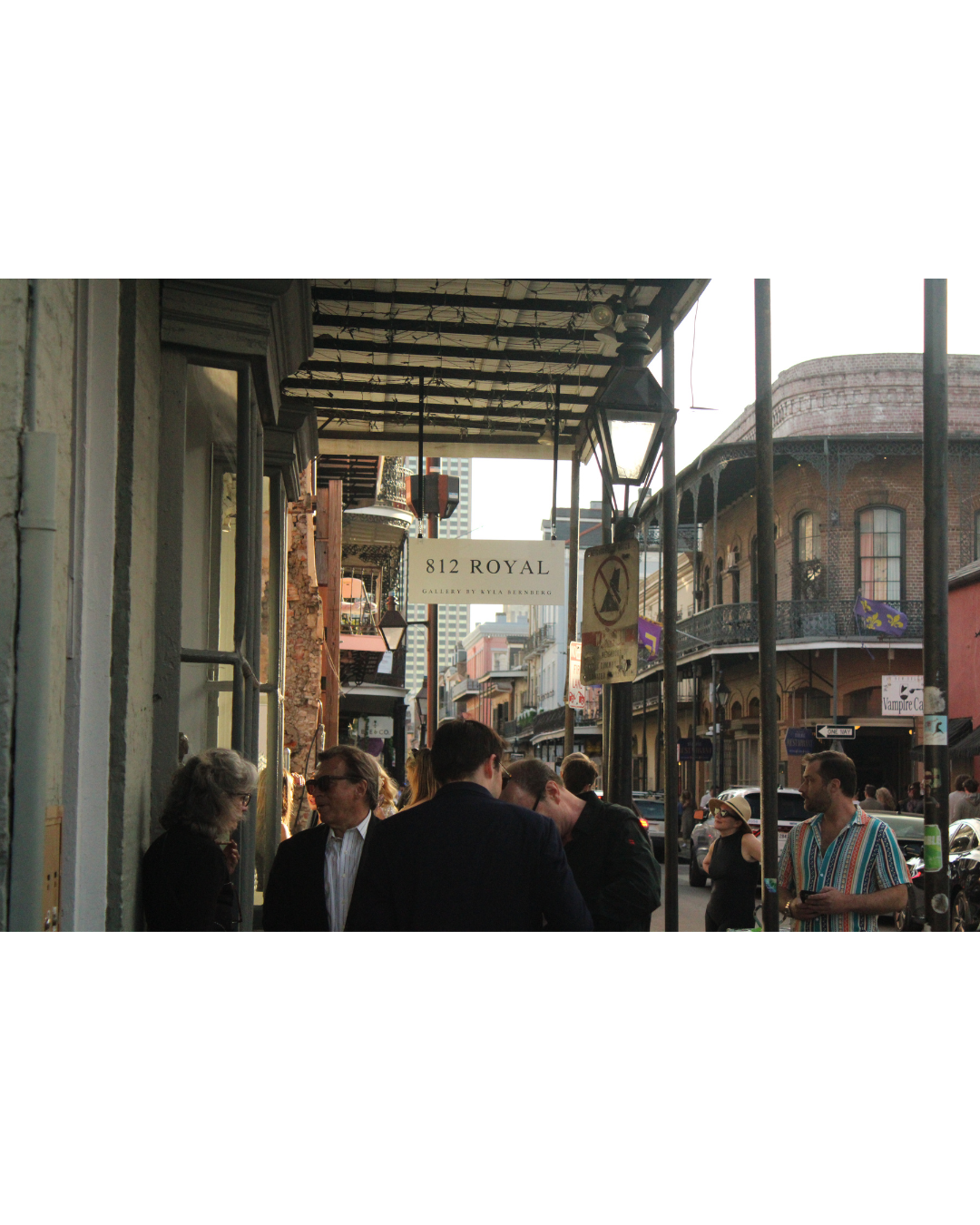 People standing and talking on a busy city street with a sign that reads '812 Royal Gallery by Kyla Seberberg' and historic buildings in the background.