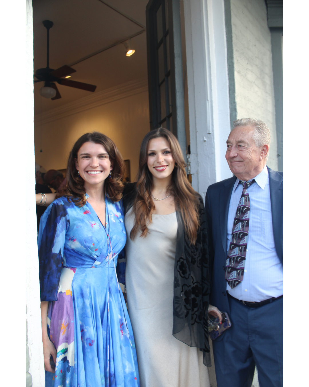 Three people, two women and one man, standing together at a social gathering or event, smiling and looking at the camera, outside a building with an open door.