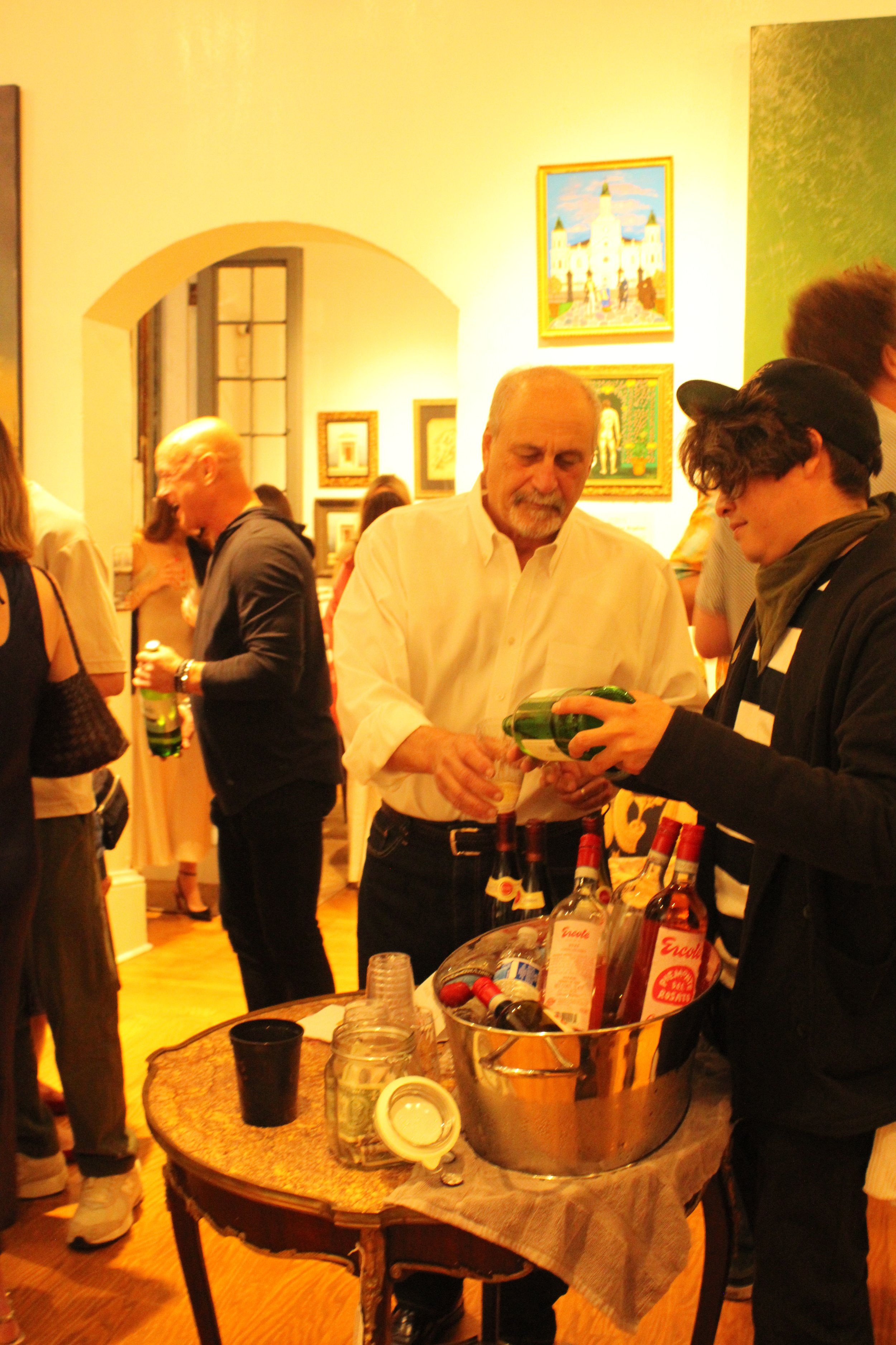People at an art opening around a table with bottles of wine, in an art gallery or museum with framed artwork on the wall.