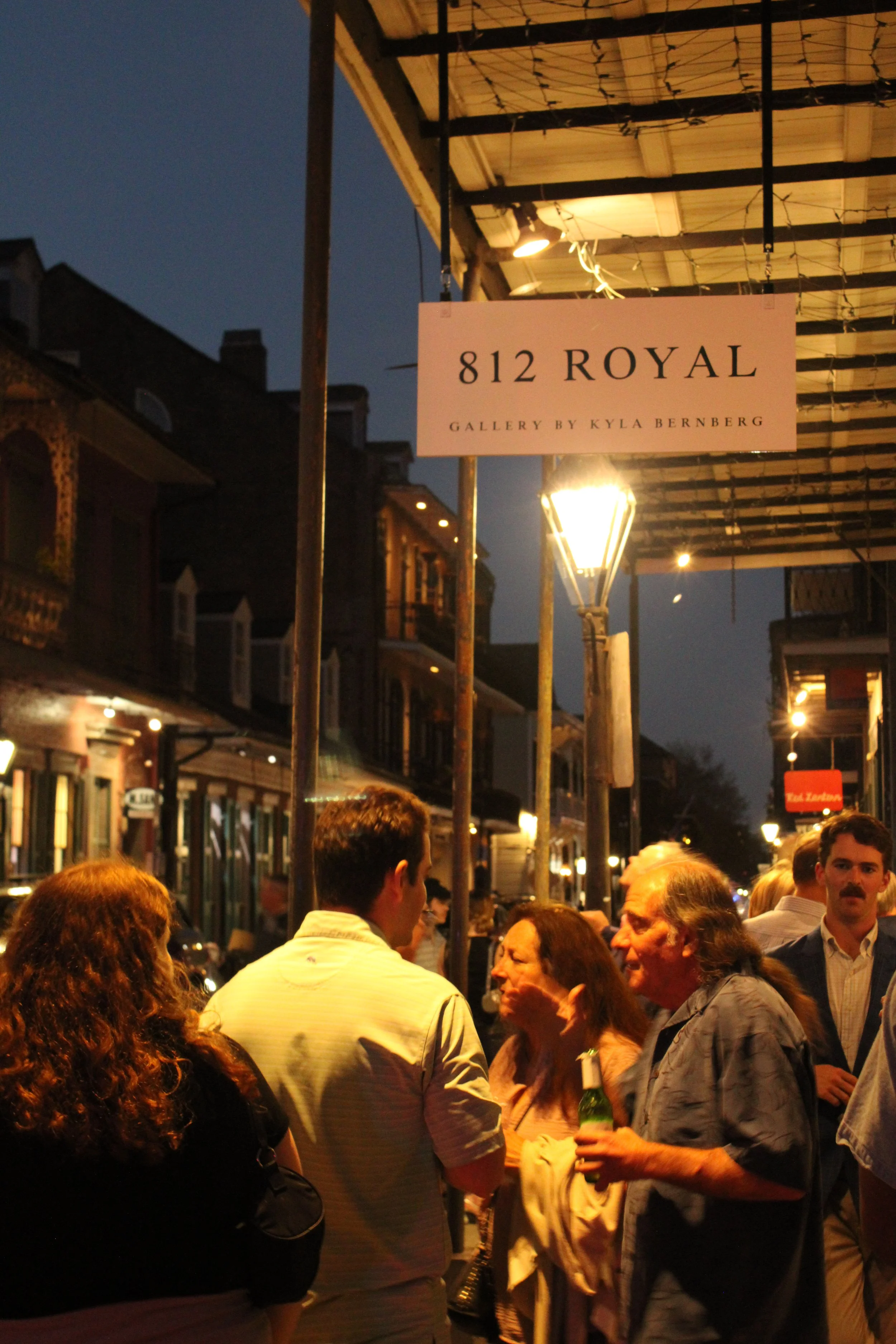 Nighttime street scene with a crowd of people gathered outside a venue with a sign that reads '812 Royal' and 'Gallery by Kyla Bernberg', illuminated by warm streetlights and decorative string lights.