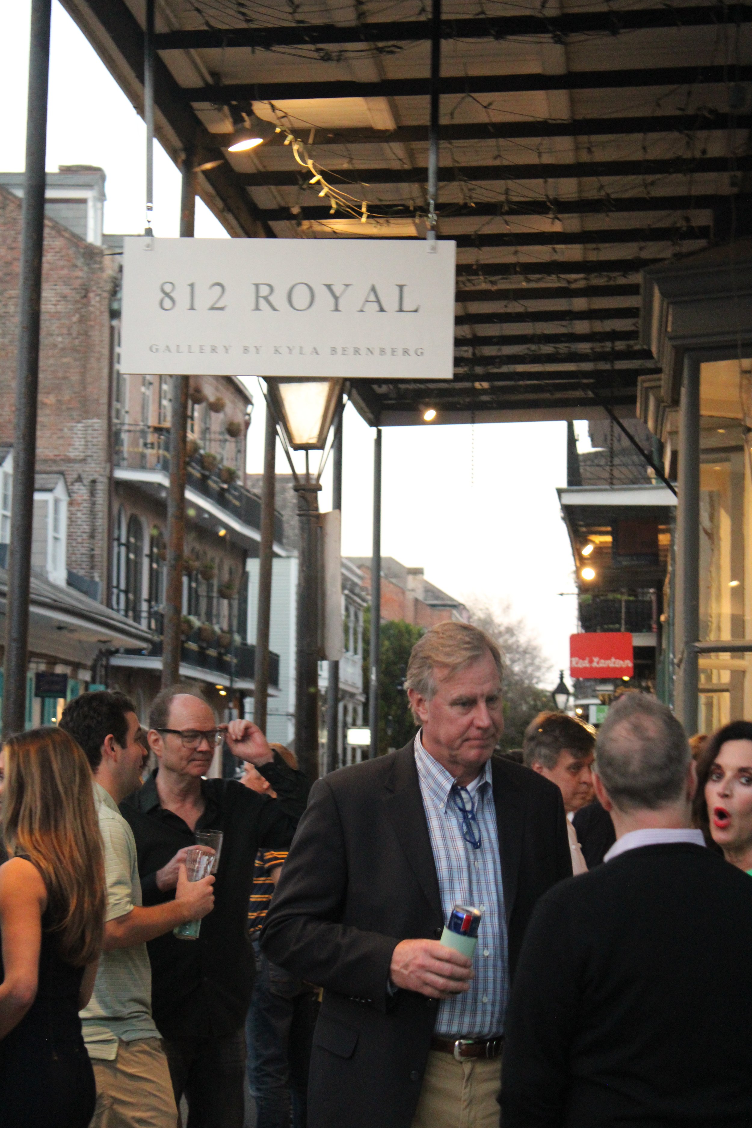 People dressed in business casual clothing gathered outdoors under a building awning, with a sign reading '812 Royal' and business names below. The scene appears to be at an event or gathering in an urban area.