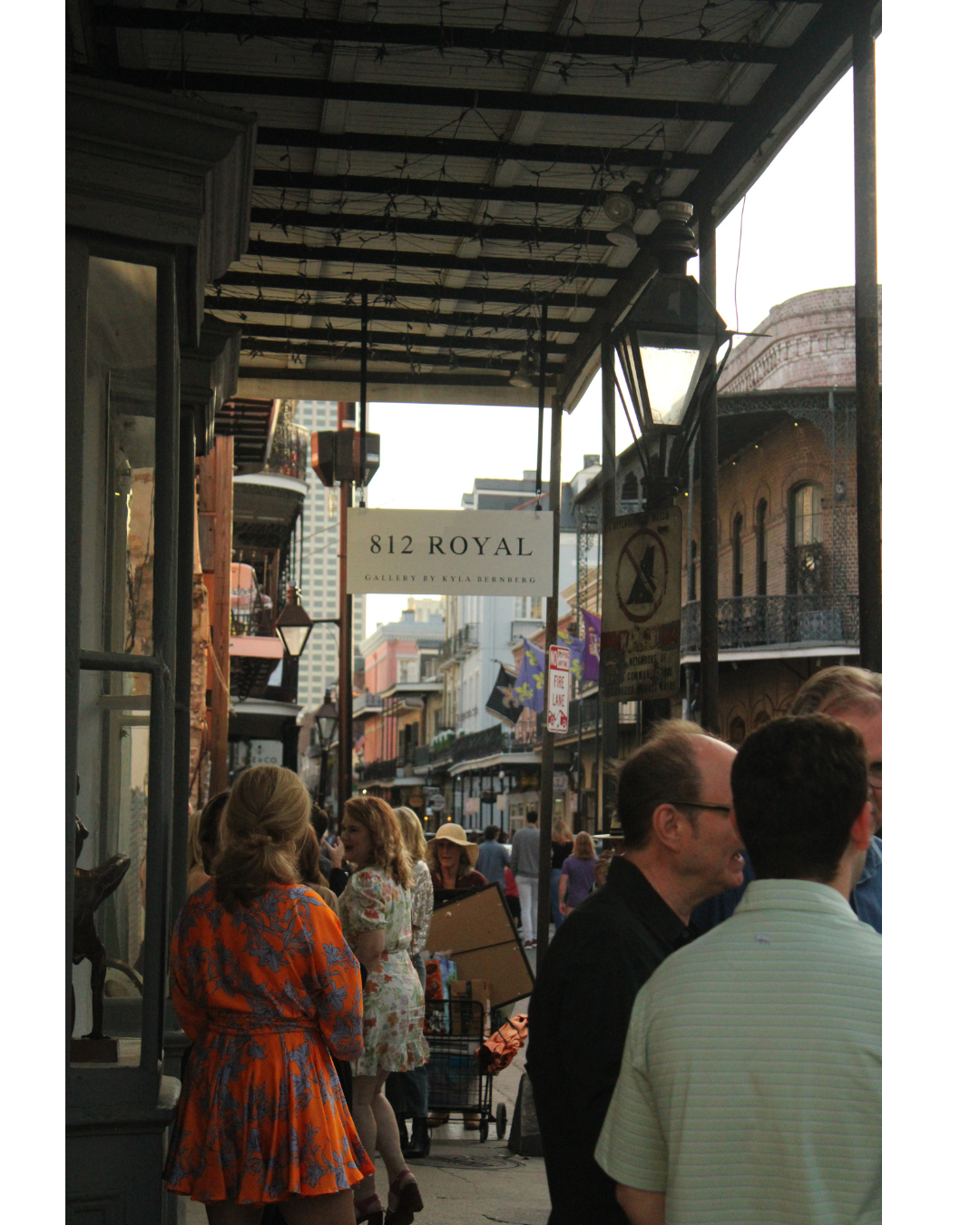 People walking and standing on a busy city street with shops, buildings, and signs, including a street sign for 812 Royal and a sign indicating a gallery by Kyla Bernberg. It appears to be late afternoon or early evening.