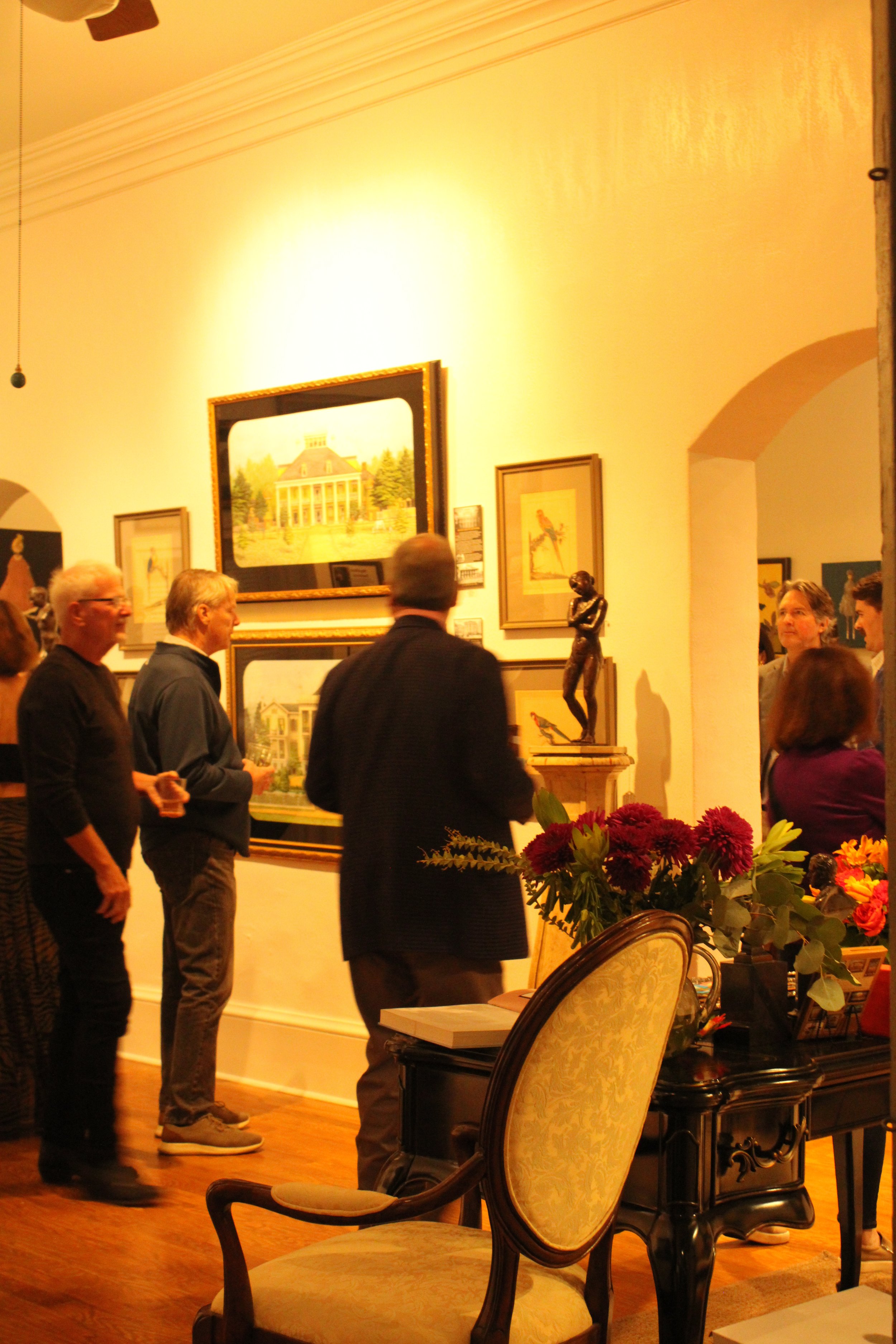 People standing and chatting inside an art gallery with framed paintings on the wall, a large central painting of a house, and a sculpture of a woman on a pedestal. There are flowers on a table in the foreground and a floral-patterned chair. The room