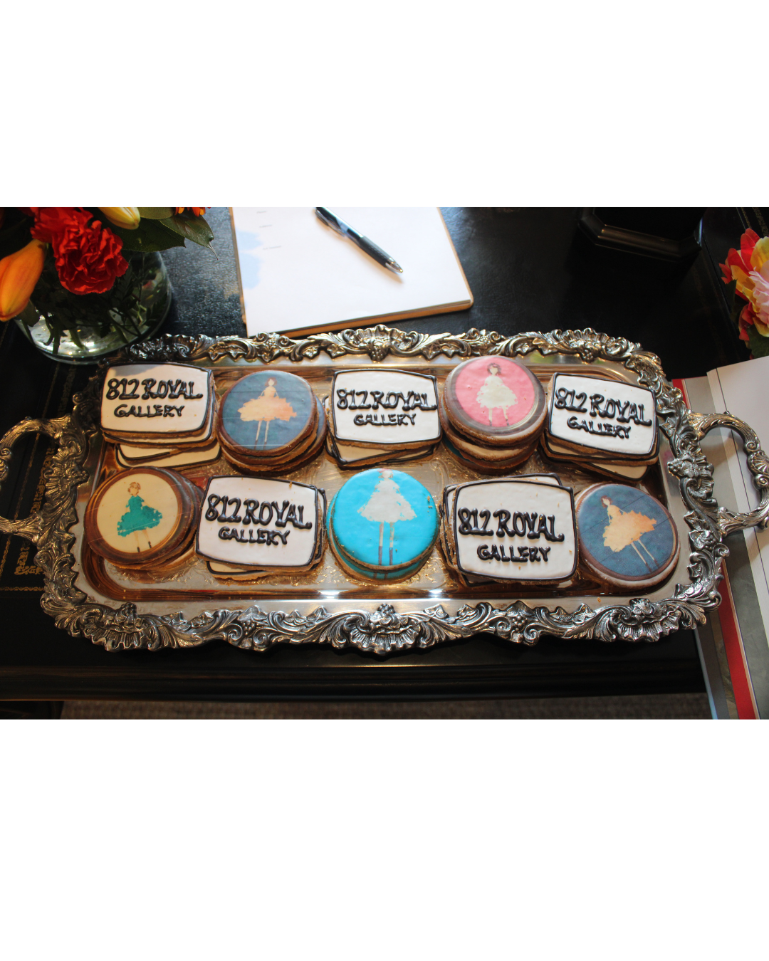 Tray of decorated cookies with the text '812 Royal Gallery' and ballerina silhouettes in pink, blue, and beige colors, placed on a black table with flowers, a notepad, and a pen.