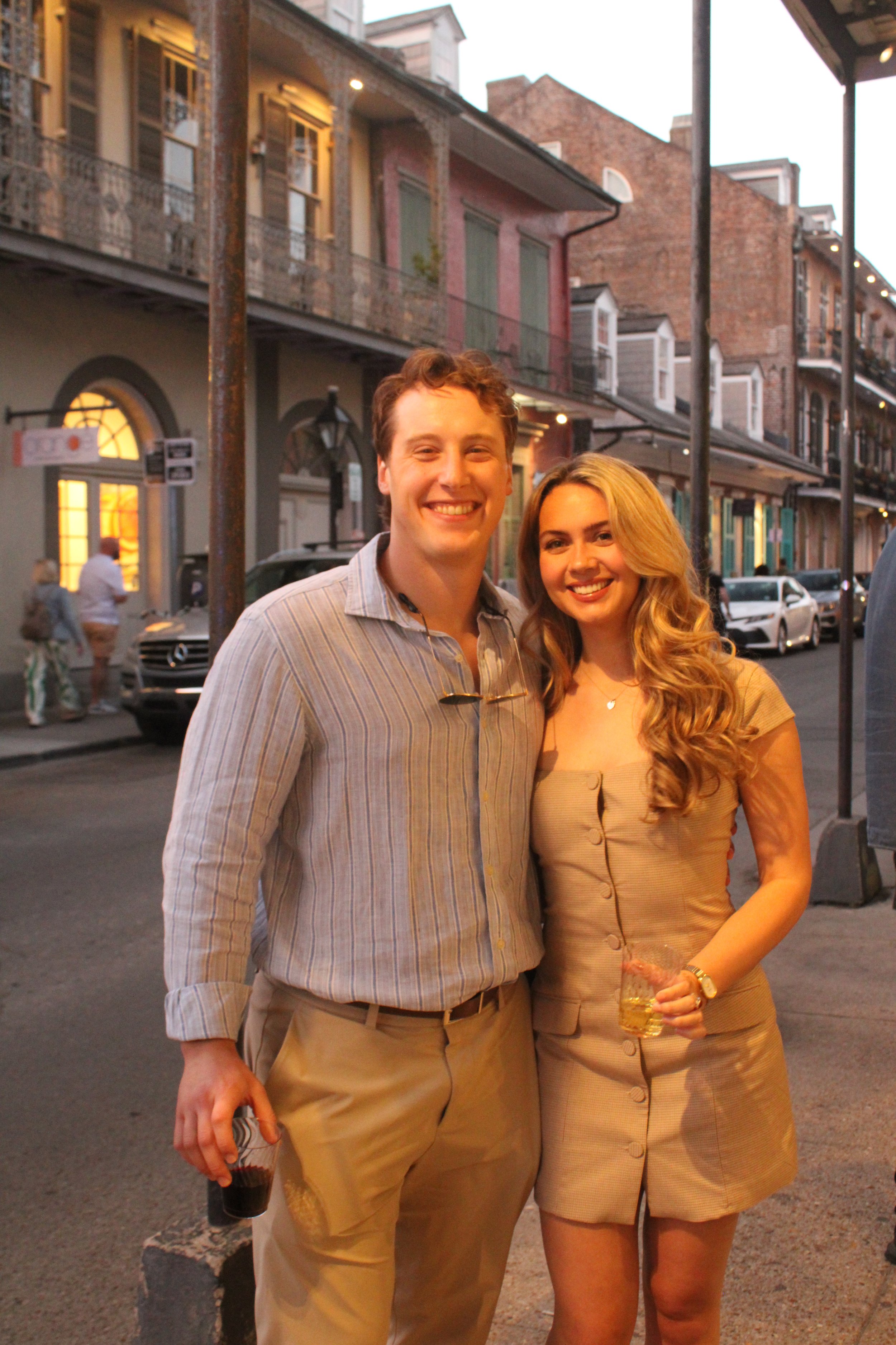 A smiling man and woman posing together on a city street during evening hours. The man is holding a drink, wearing a striped shirt and beige pants. The woman is holding a glass with a beverage, wearing a beige dress with buttons. There are cars parke