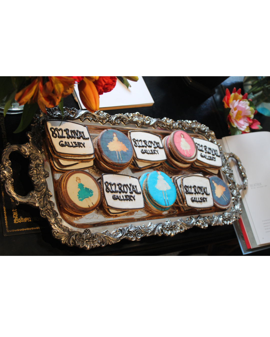 Decorative cookies with images of ballerinas and the words 'Ballerina Gallery' on a silver tray, surrounded by flowers and notebooks.