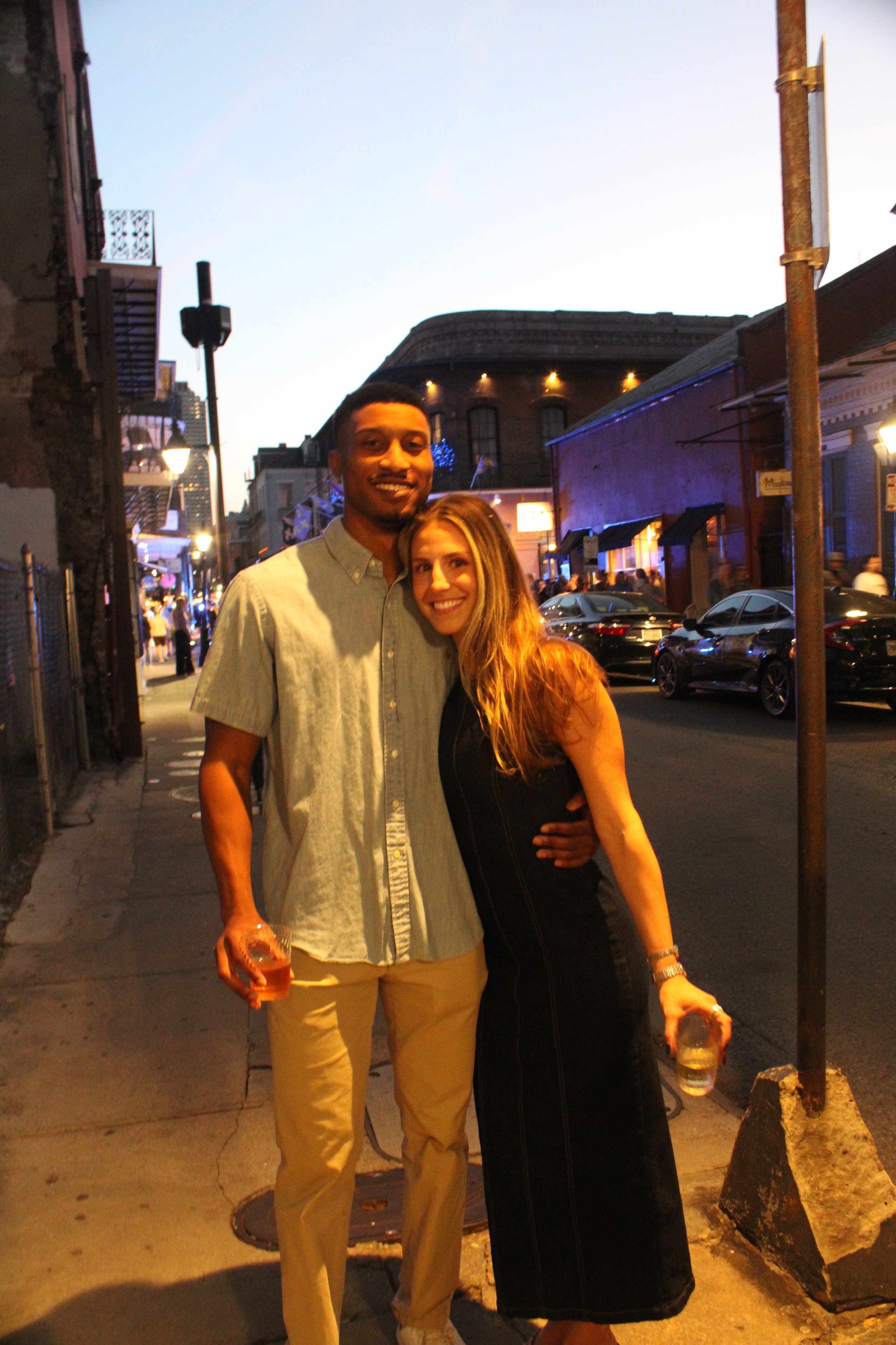 A smiling young man and woman hugging on a city sidewalk during the evening. The man is holding a drink and the woman is holding a drink in a plastic cup. There are parked cars and buildings with illuminated windows in the background.
